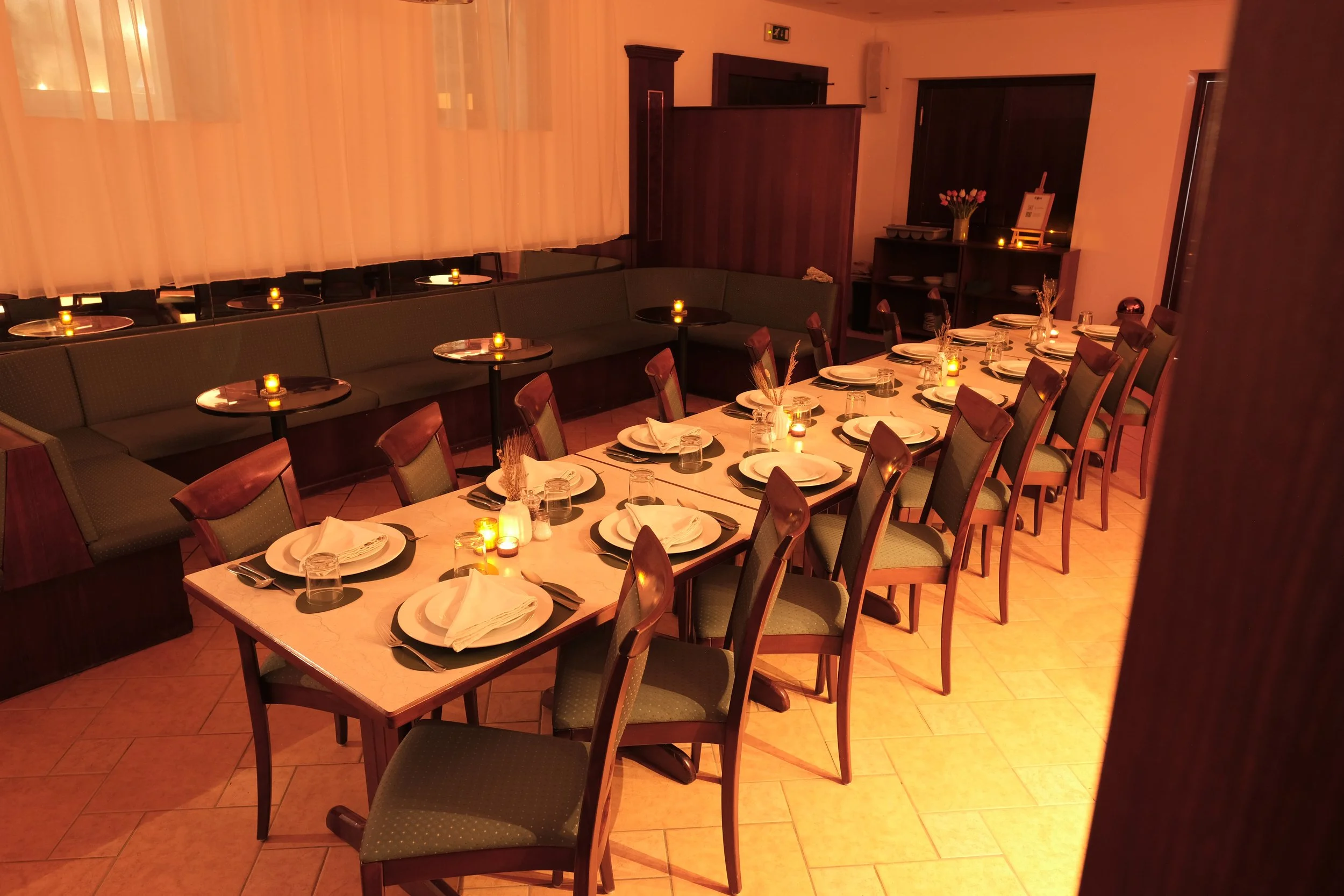 Empty restaurant dining room with a long table set for a meal, surrounded by wooden chairs, candlelit ambiance, and pastel pink lighting.