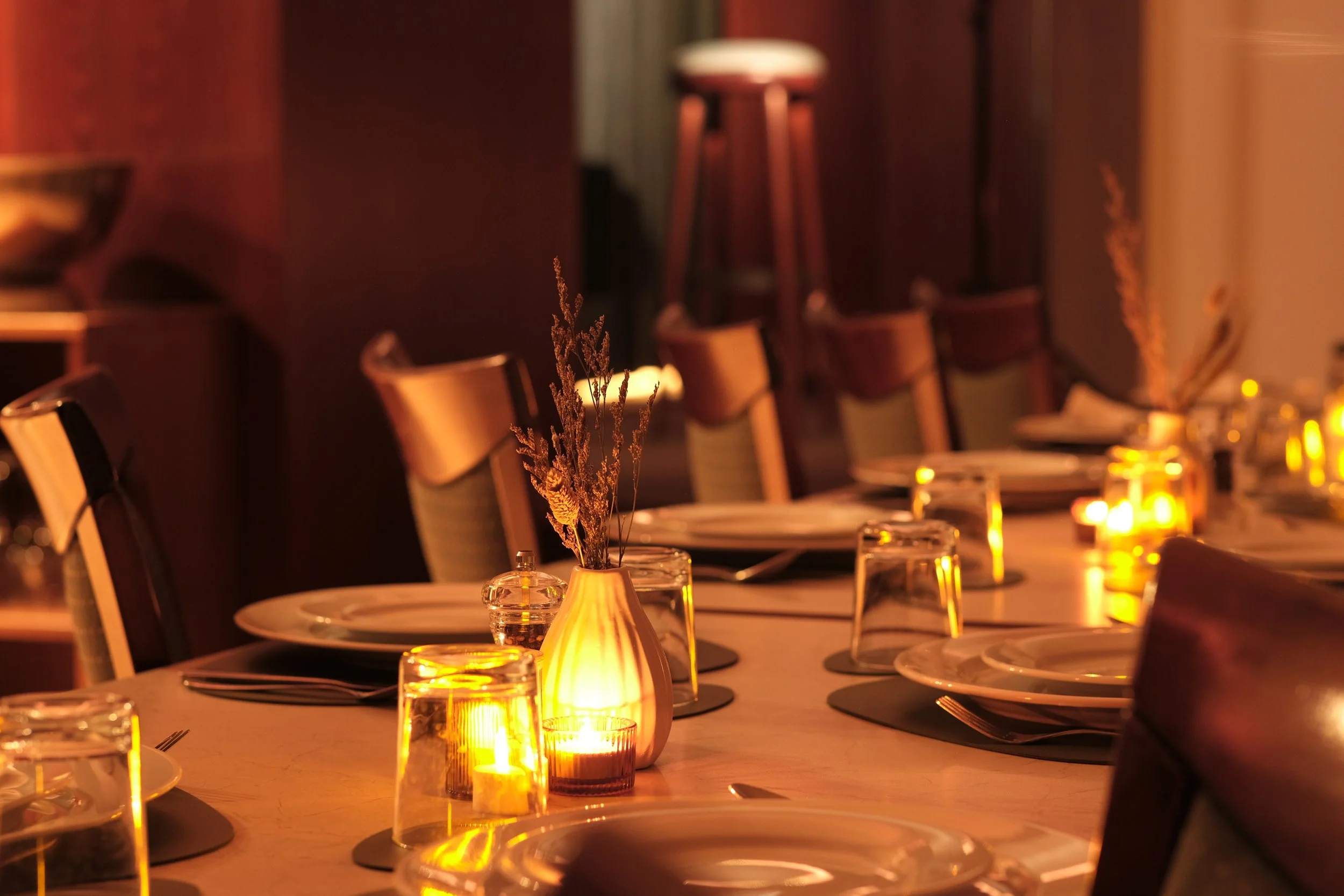 A dimly lit dining table set with plates, glasses, candles, and small vases with dried flowers, creating an elegant and cozy ambiance.