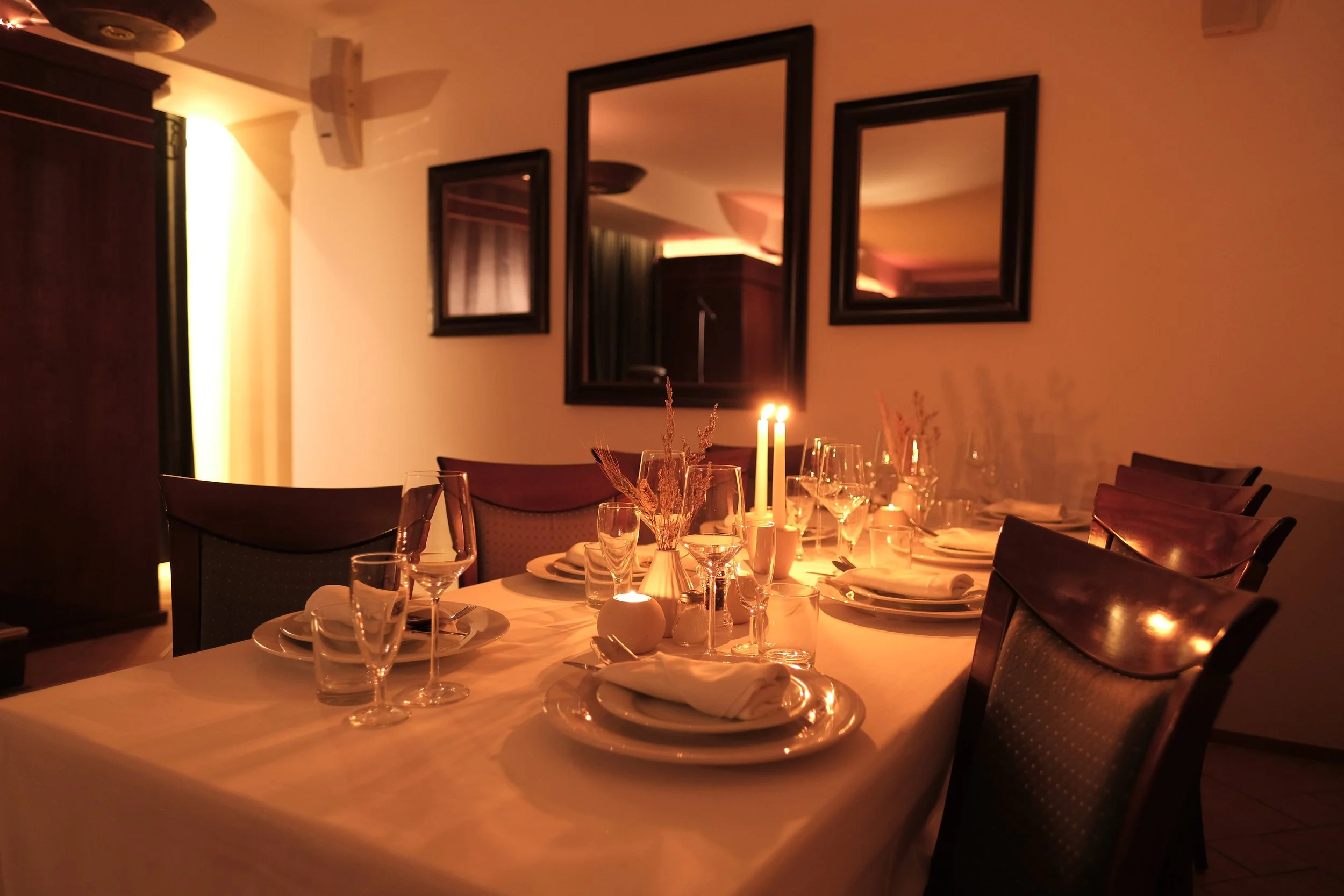 A dimly lit dining room with a table set for a meal, featuring candles, wine glasses, plates, and silverware, with three mirrors on the wall reflecting the warm lighting.
