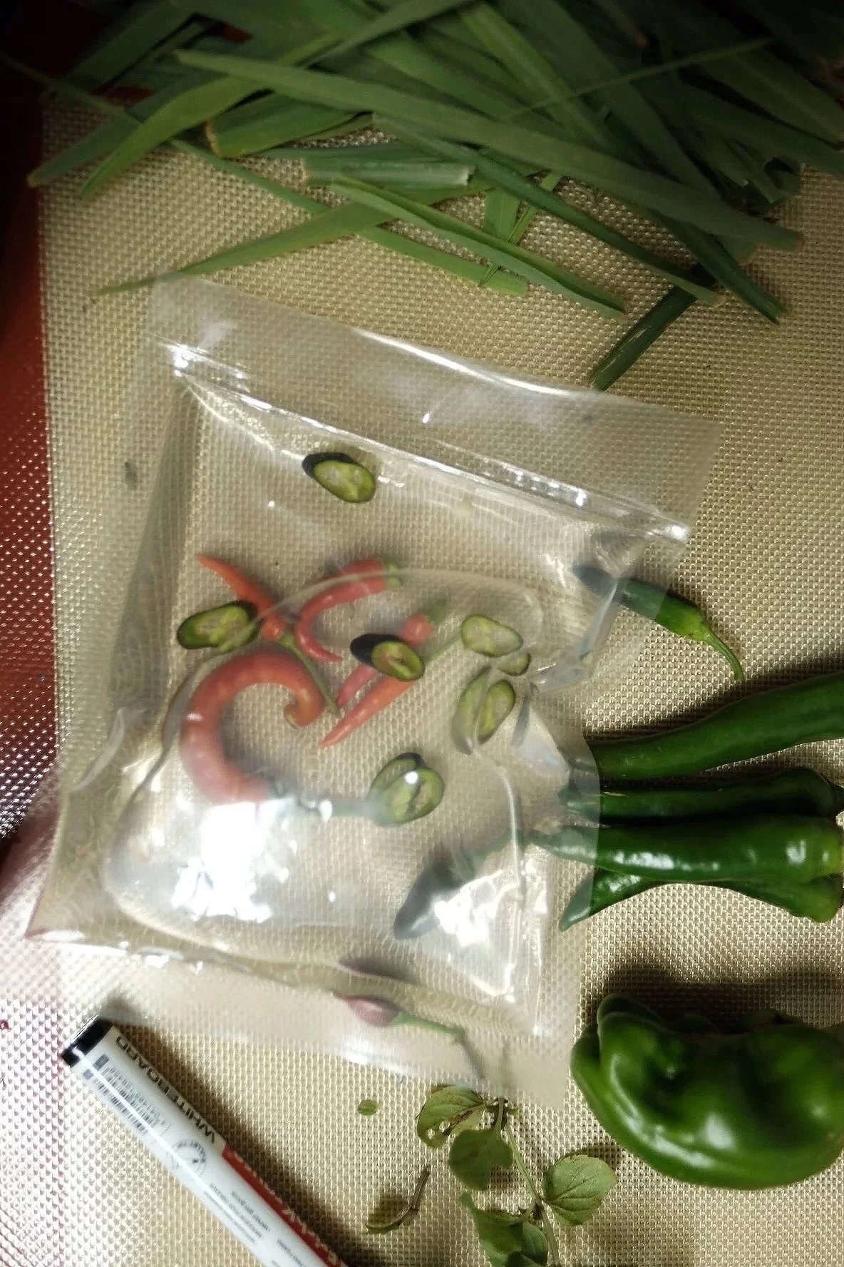 Green chili peppers, jalapeño pepper slices in a plastic bag, and other vegetables on a beige surface.