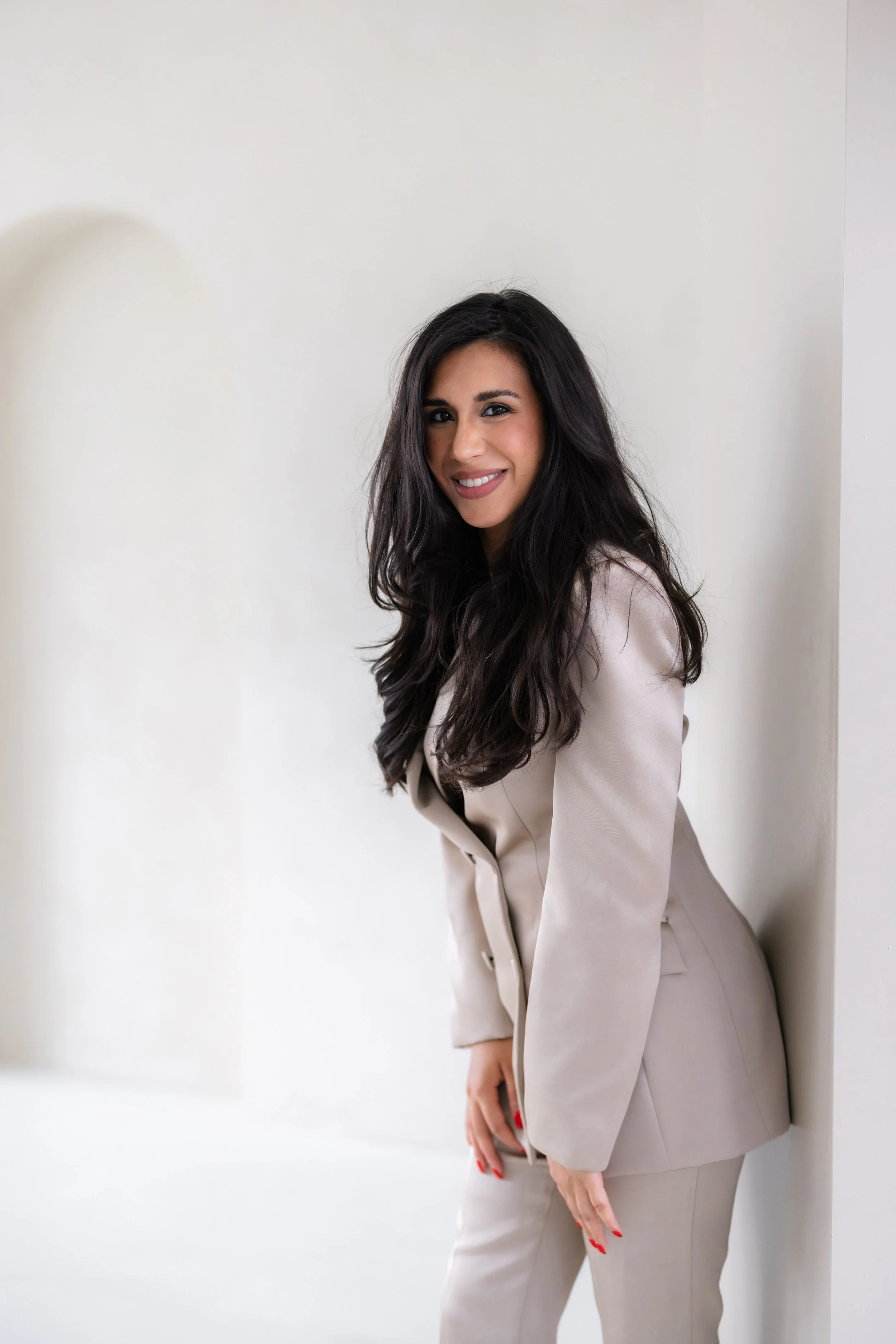 A woman with long dark hair, dressed in a beige suit, leaning against a white wall with a soft smile.
