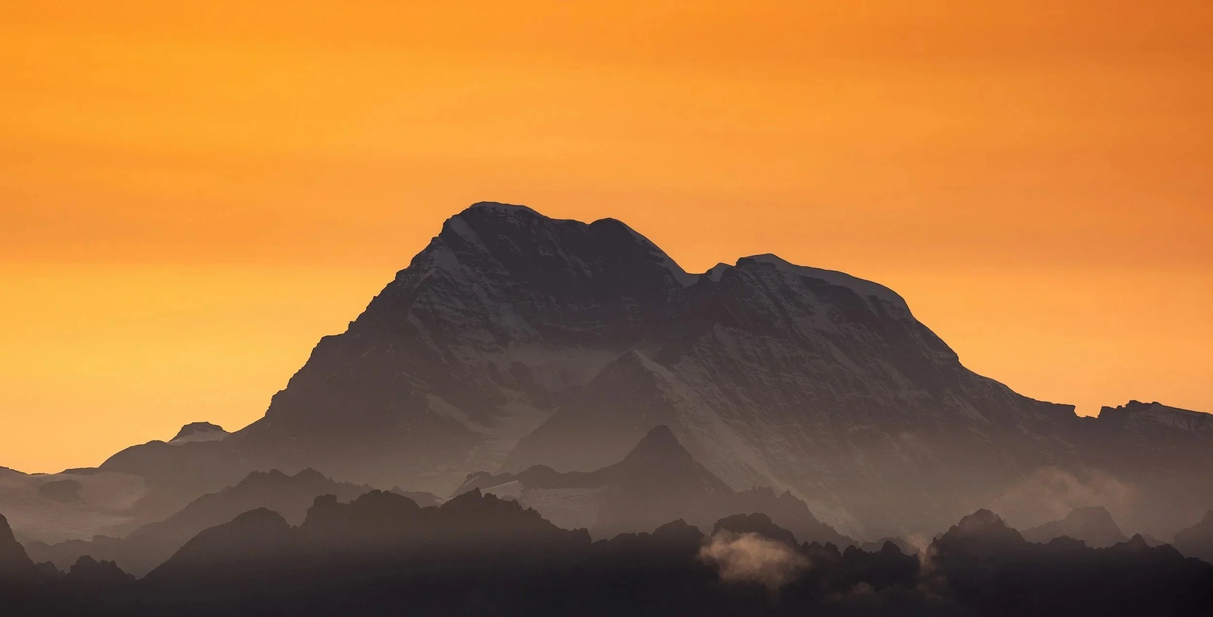 Mountain silhouette against a sunset sky with orange hues and some clouds near the base of the mountain.