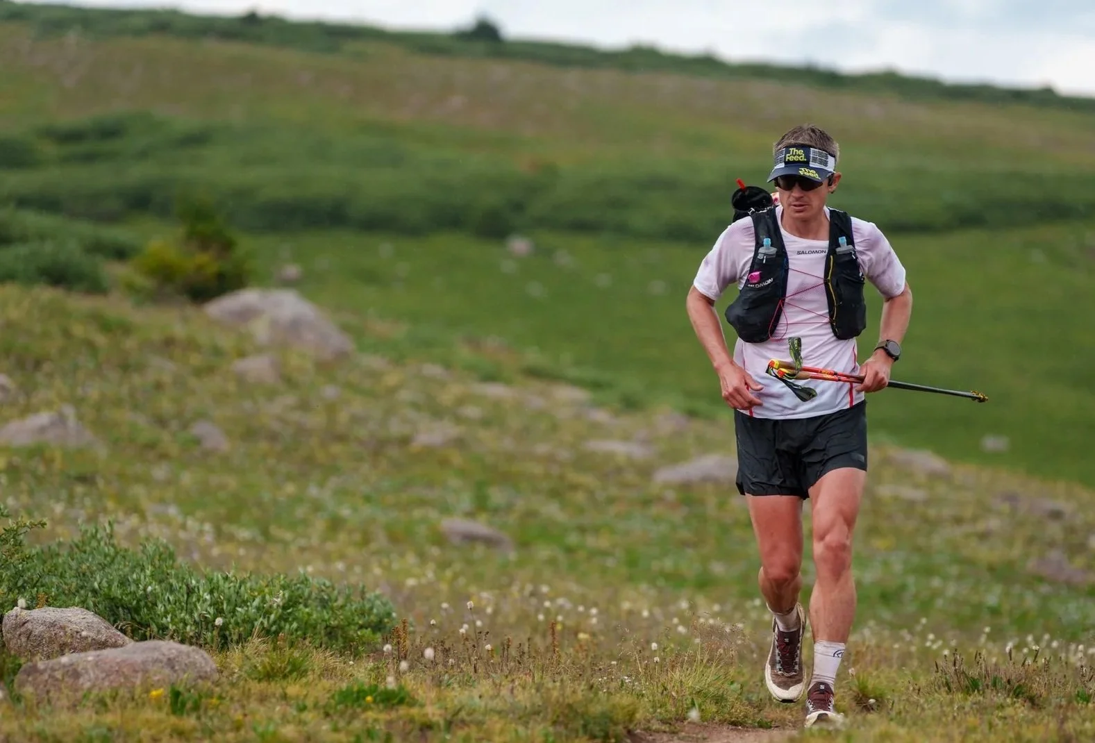 Male trail runner wearing a white shirt, black shorts, and sunglasses running on a dirt trail in a grassy, hilly landscape with rocks and green vegetation.
