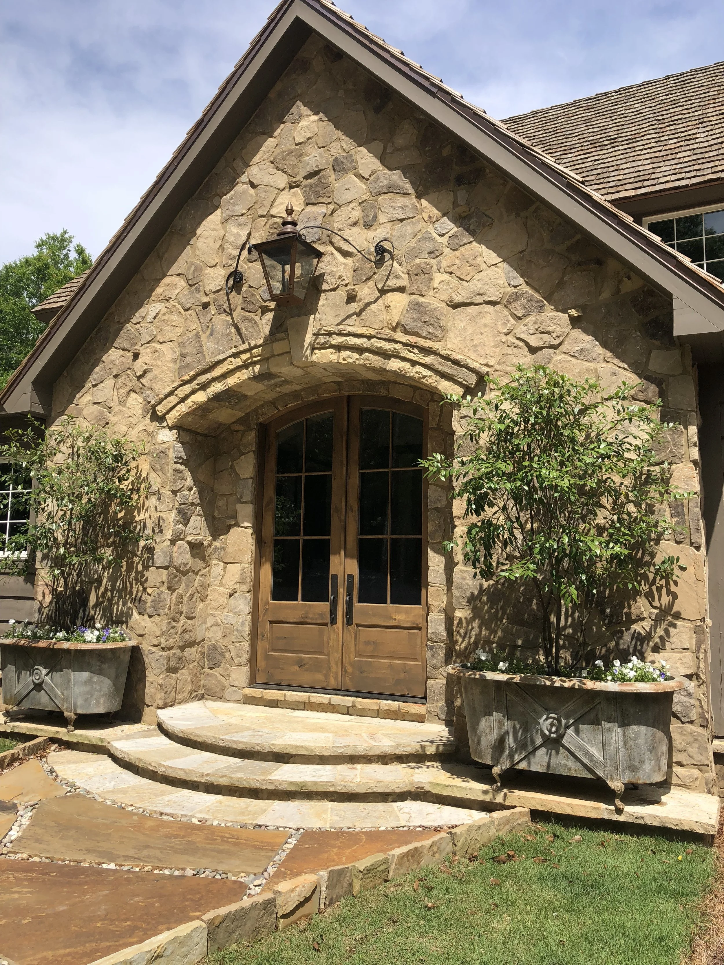Front entrance of a house with stone facade, wooden double doors, potted plants on either side, and a hanging lantern light above the door.