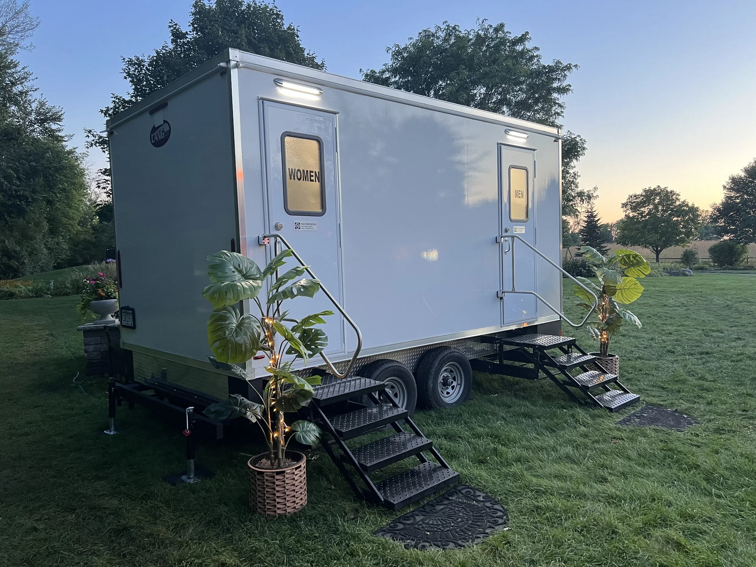 mobile restroom trailer outside in the grass at dusk with a potted plant to the left