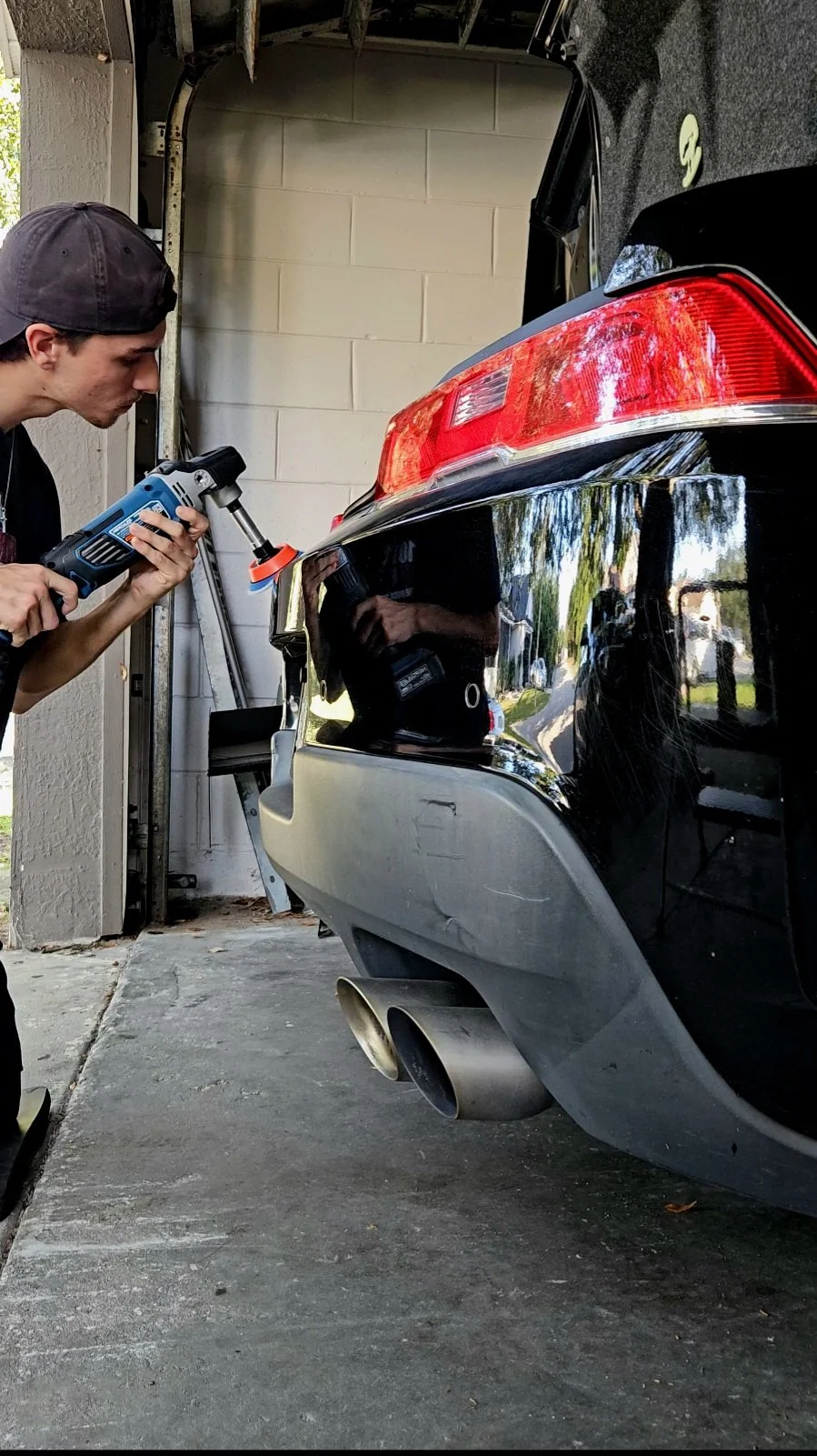A person using a power polisher on the rear bumper of a black sports car in a garage.