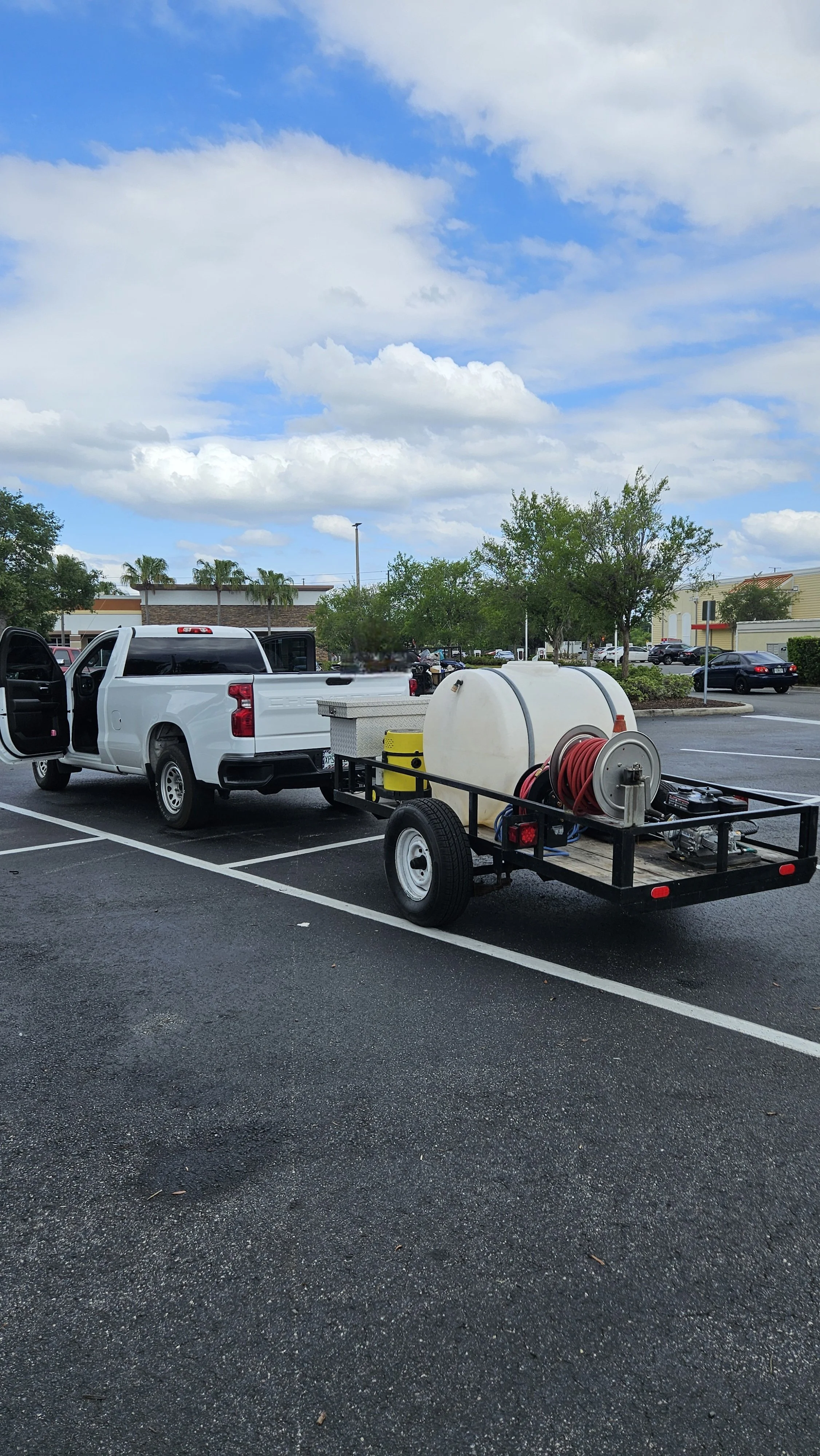 A white pickup truck towing a trailer with a large water tank, hoses, and equipment in a parking lot surrounded by trees and buildings under a partly cloudy sky.