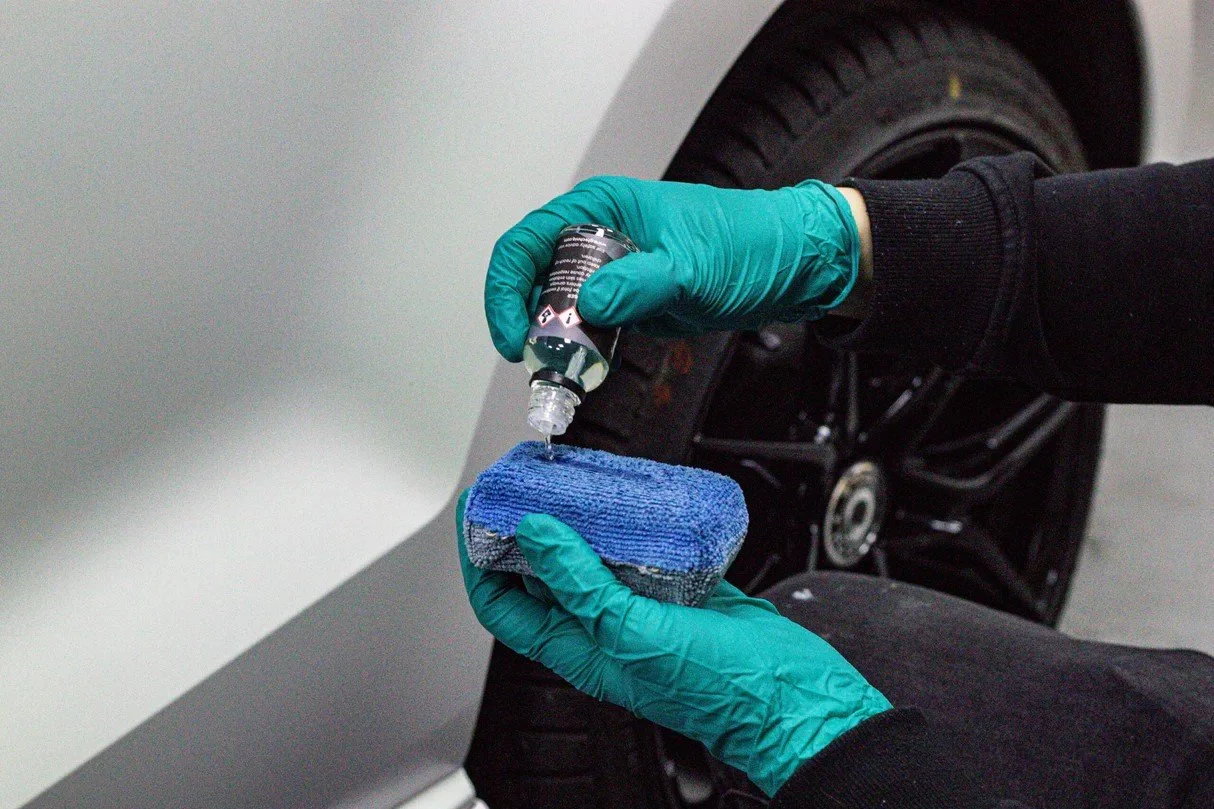 Person wearing teal gloves cleaning white car with a spray bottle and blue cloth near a black car wheel.