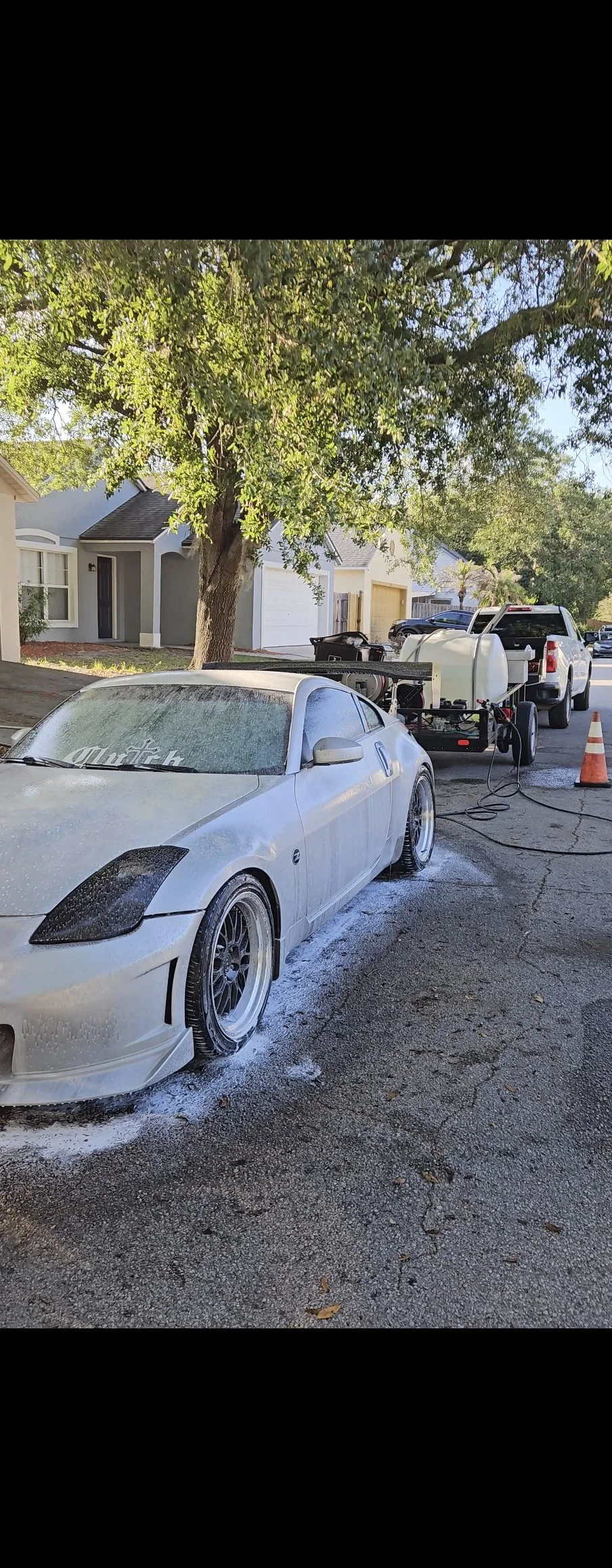 A white sports car is being washed with water and soap. A foam cannon is attached to a pressure washer, and the water is spraying onto the car. The car is parked on a residential street with houses and trees in the background.
