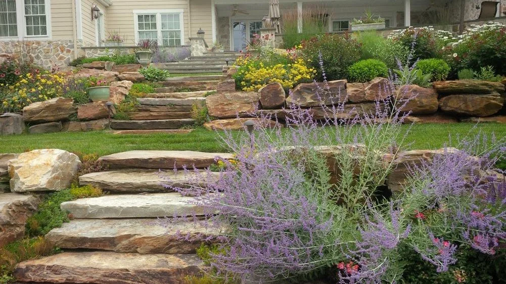 Stone steps leading up a landscaped garden with various plants and flowers, in front of a house with a porch.