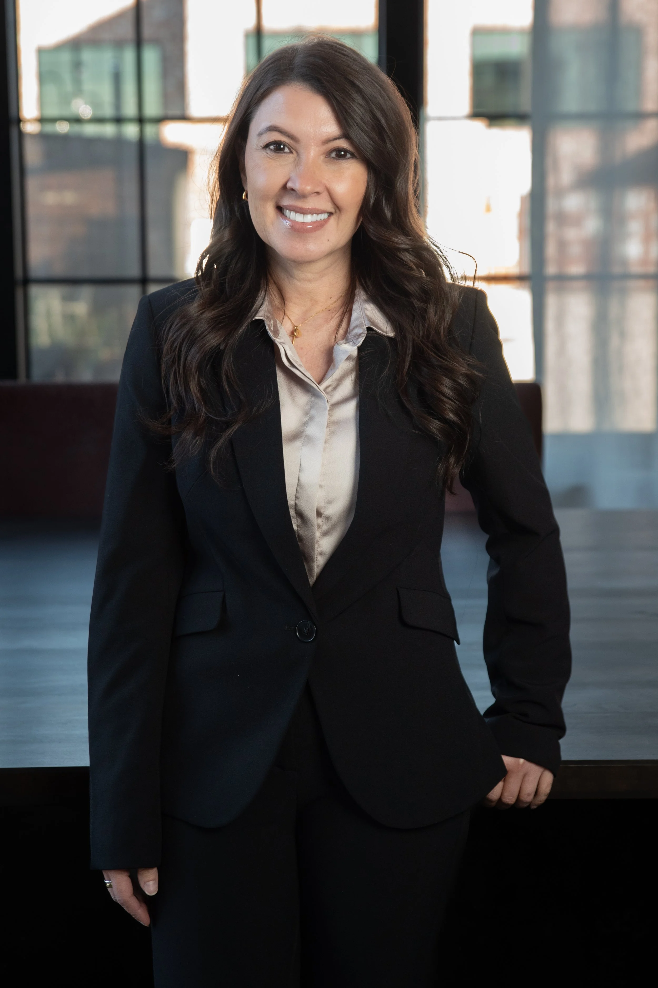 A woman in a black business suit standing in a modern office with large windows behind her.