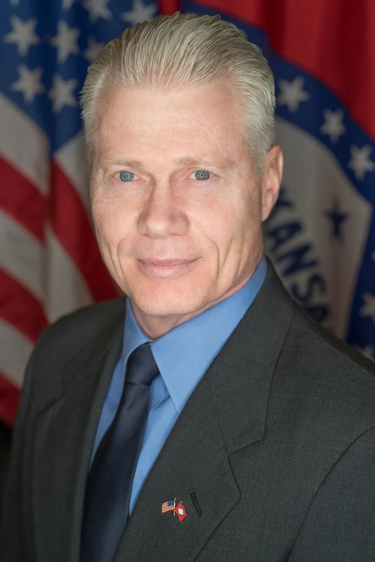 Portrait of a man in a suit with an American flag badge, standing in front of a blurred American flag background. Headshot of politician running for office.