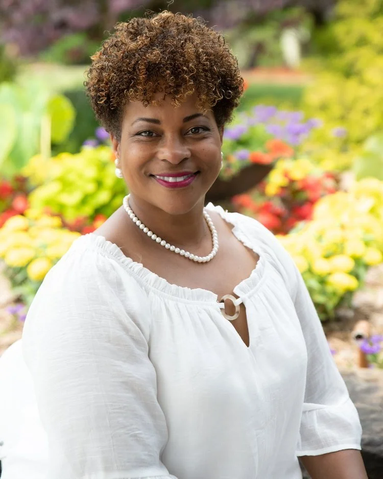 Smiling woman with short curly hair wearing a white blouse and pearl necklace, standing outdoors in front of colorful flowers.