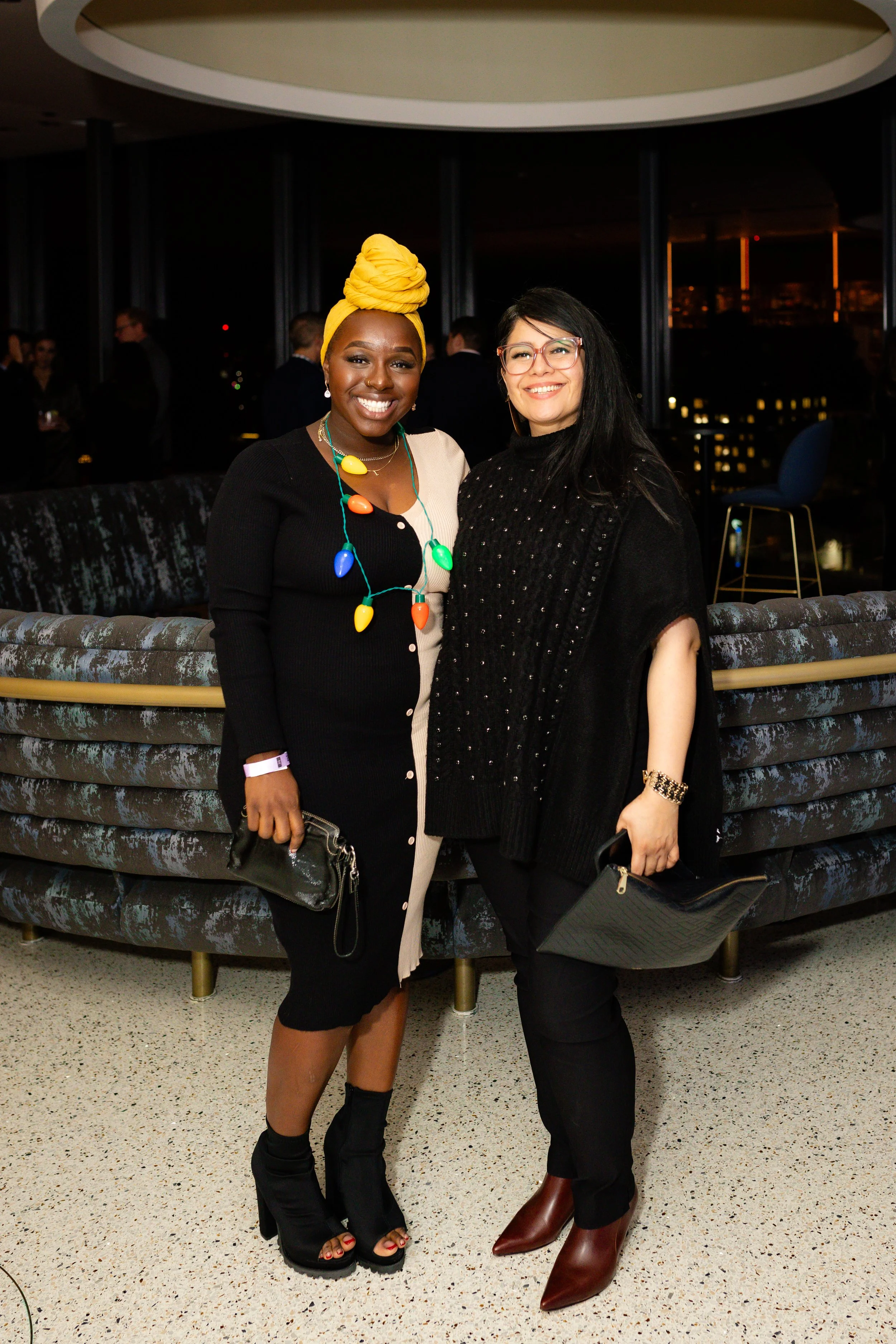 Two women standing together smiling in an indoor setting at night, with city lights visible through the windows behind them.