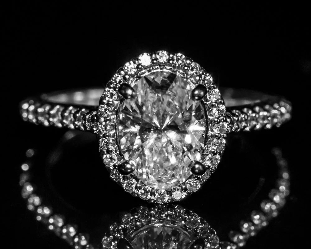 Close-up of a diamond engagement ring with a large oval-shaped center stone and a band encrusted with smaller diamonds, reflected on a black surface.