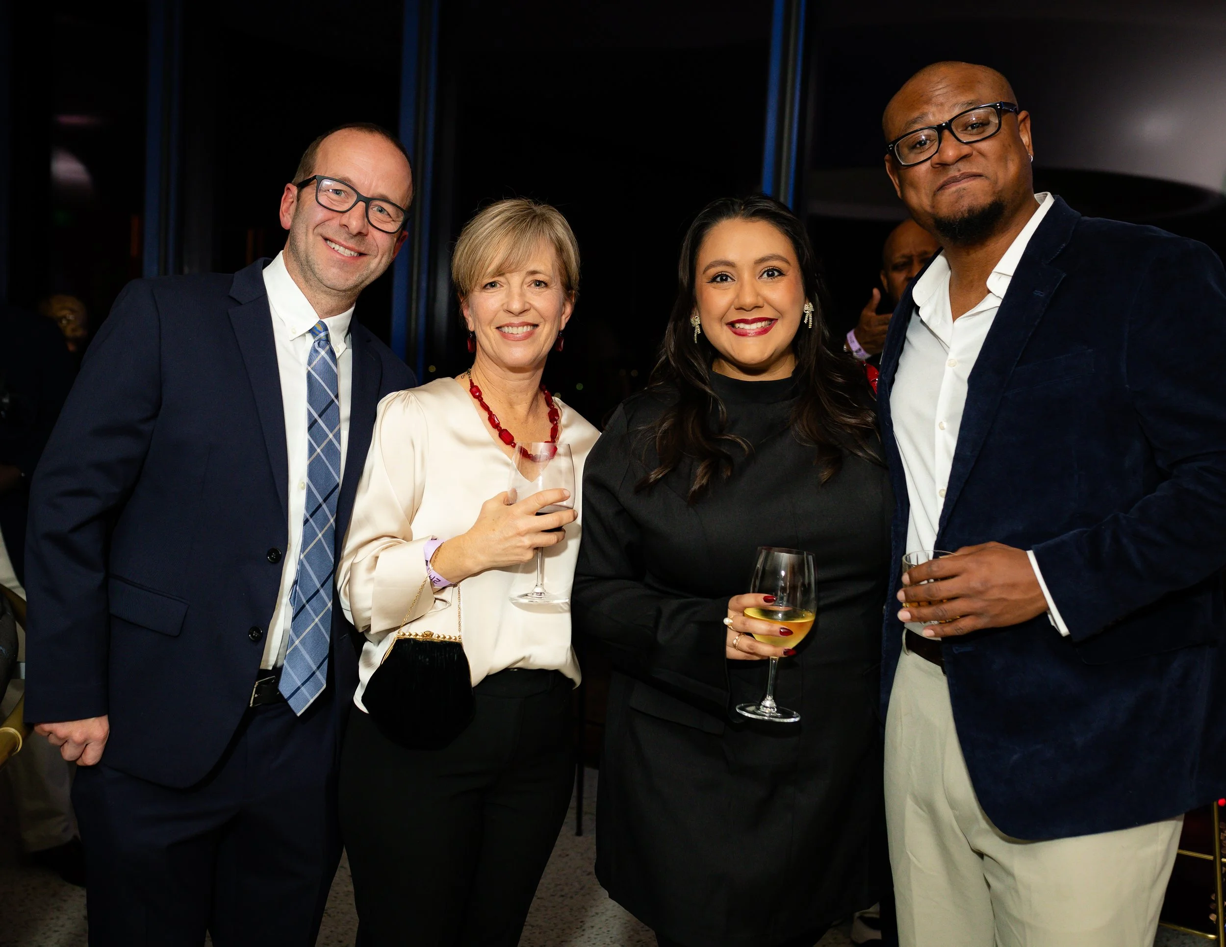 Four people at a social event, smiling and holding wine glasses, dressed in formal attire.