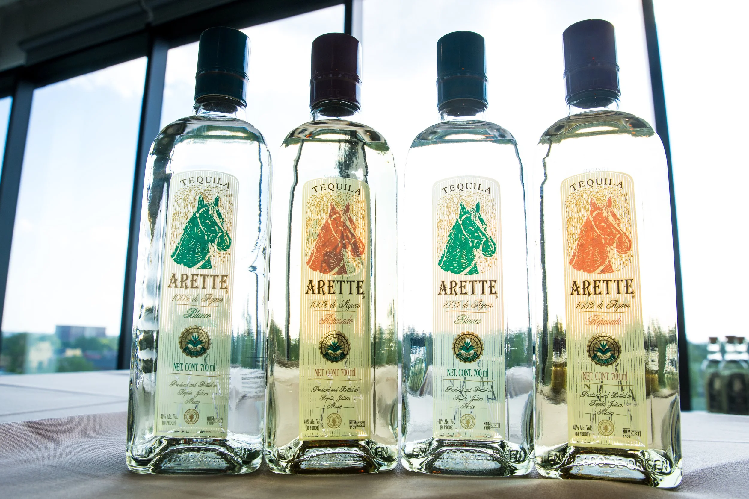Four bottles of Tequila Arete with different labels, arranged on a table. A large window with a city view is in the background.