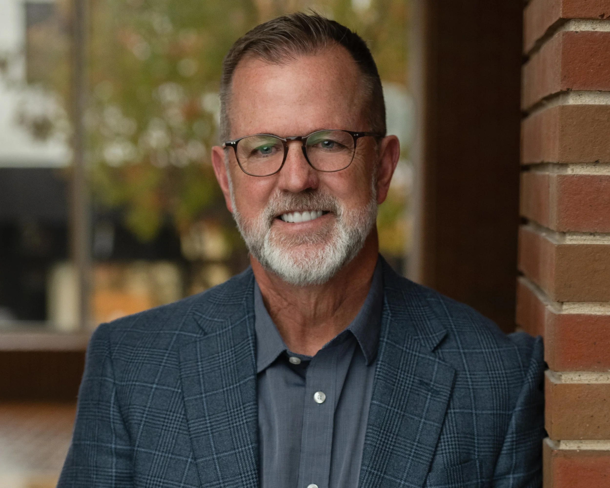 A smiling man with glasses, a beard, wearing a checkered blazer and a dark shirt, standing outdoors near a brick wall.