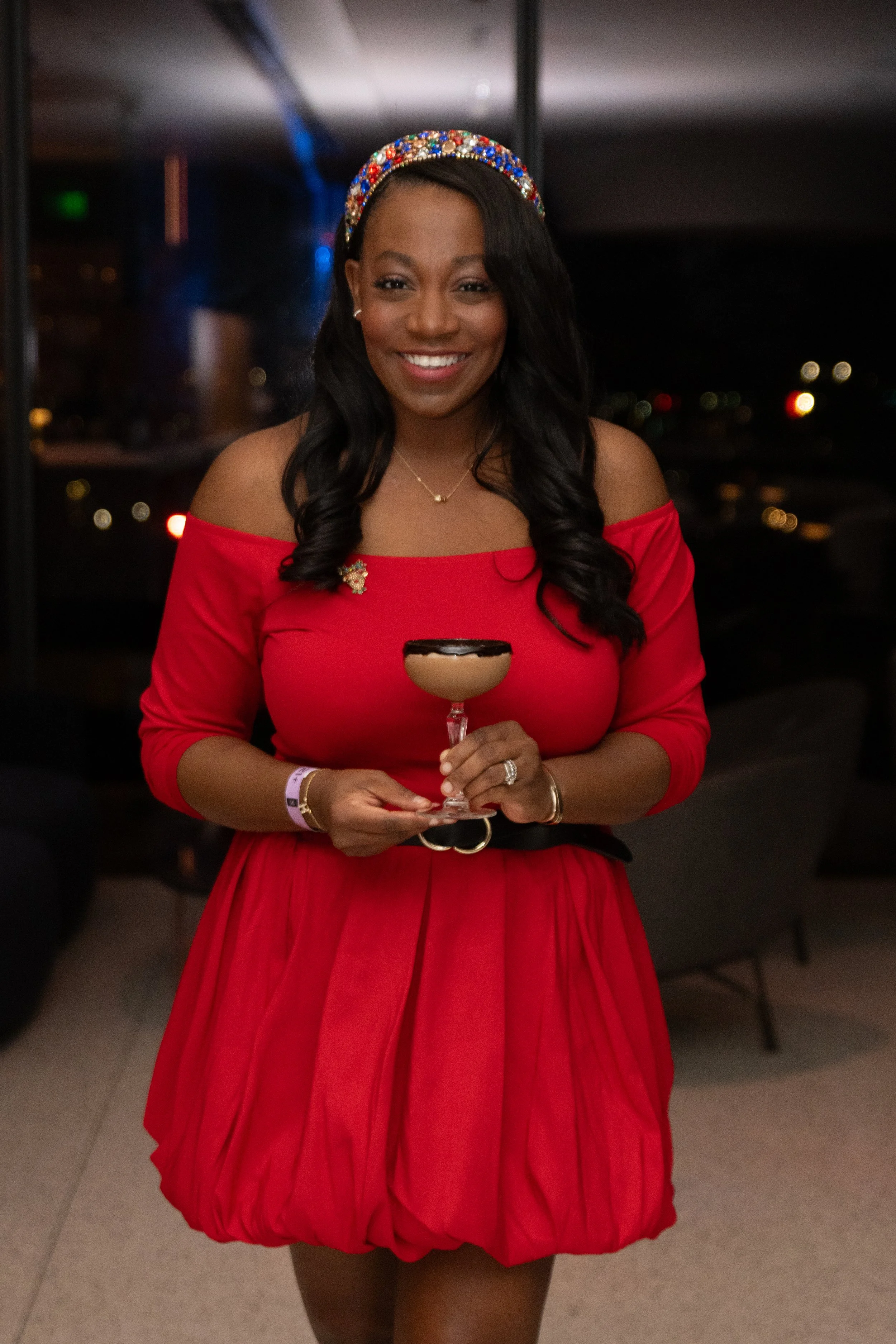 A smiling woman in a red dress with off-the-shoulder sleeves, holding a cocktail glass with a layered drink, standing in a dimly lit setting at night.