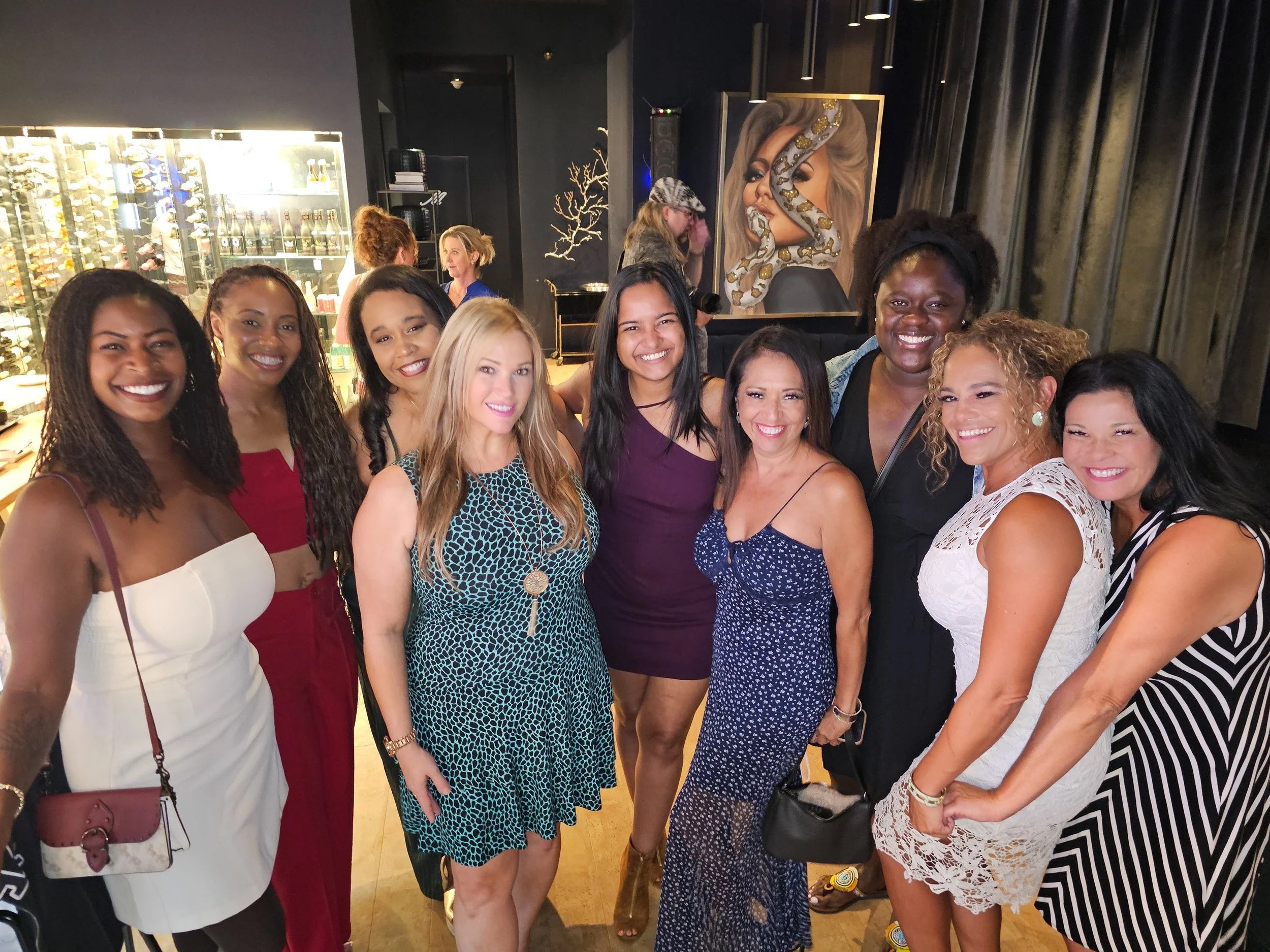 Group of ten diverse women smiling together in a social setting indoors, with artwork and a wine display in the background.