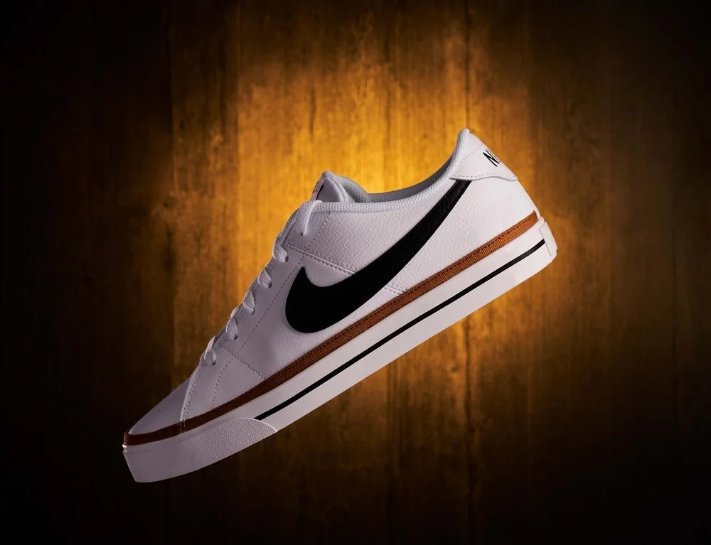 A white sneaker with a black Nike swoosh logo, brown accents, and a white midsole, floating against a wooden background.