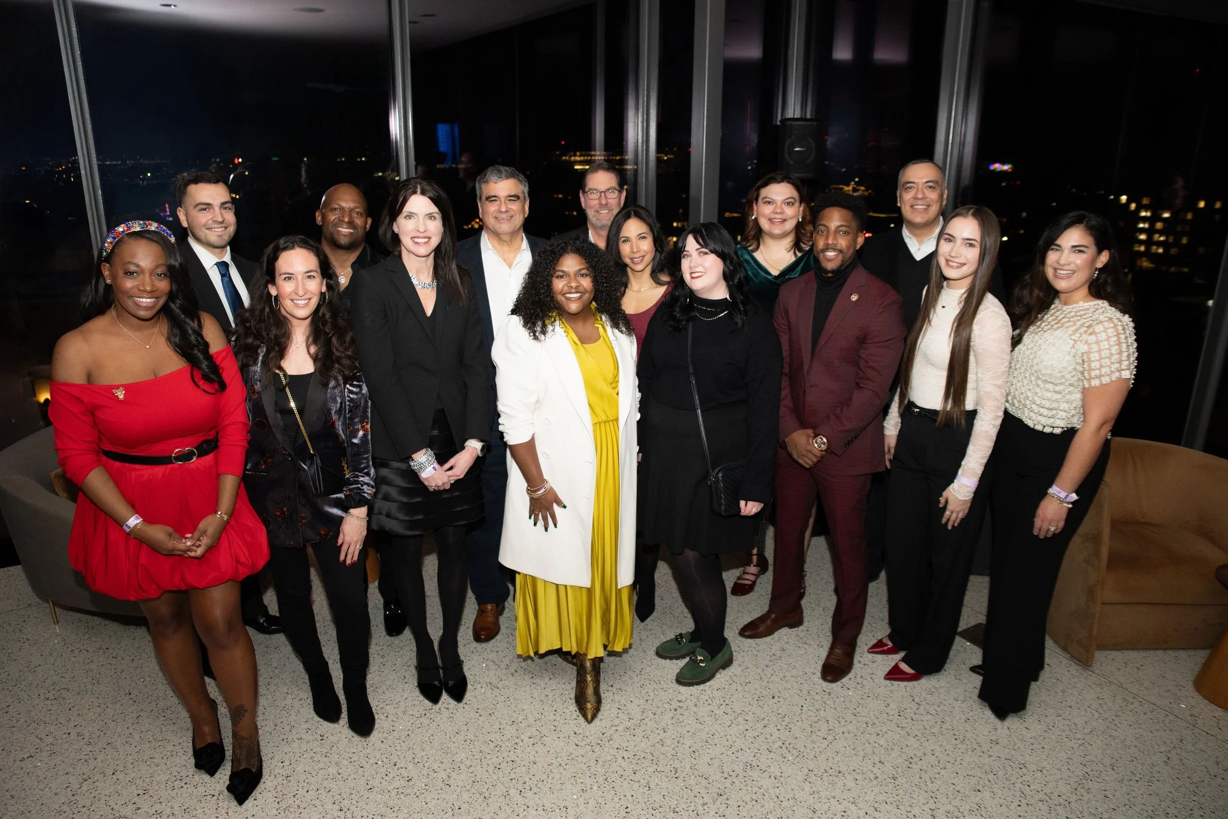 A diverse group of 16 people, dressed in formal and semi-formal attire, posing together in a high-rise building at night with city lights in the background. Break the Ice Event. Sponsors and Staff Team Photo.