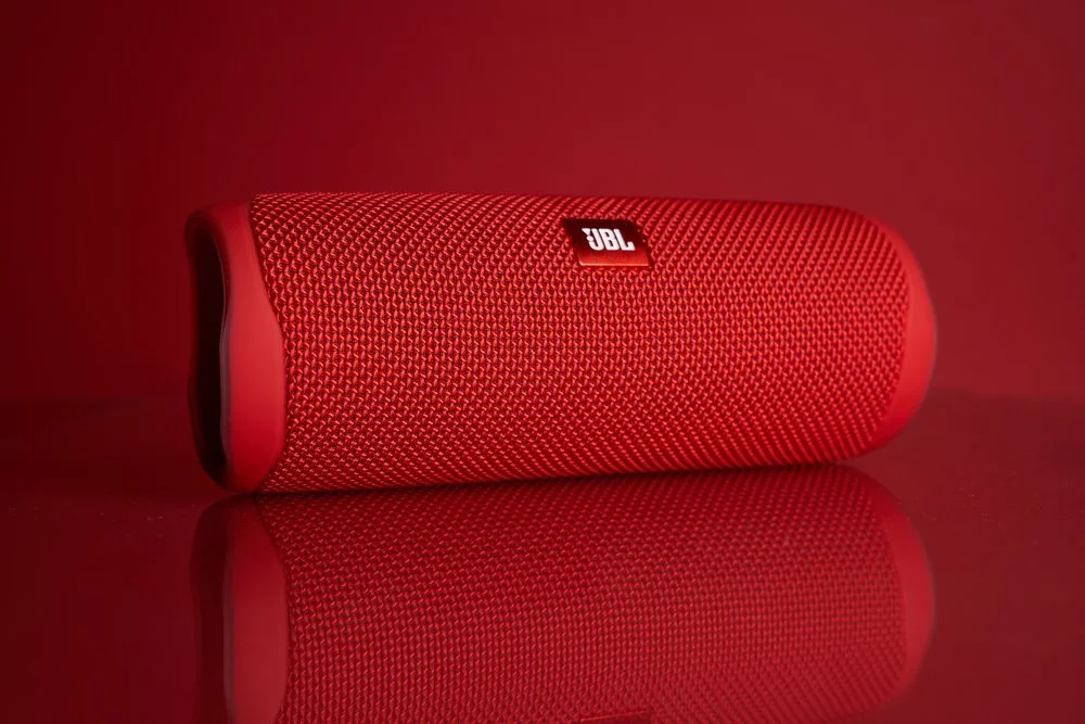 Red JBL portable Bluetooth speaker lying on a reflective surface with a red background.