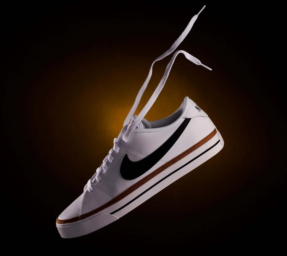 White Nike sneaker with black swoosh logo, brown and black stripe sole, suspended in mid-air against a dark background with a glowing light behind it.