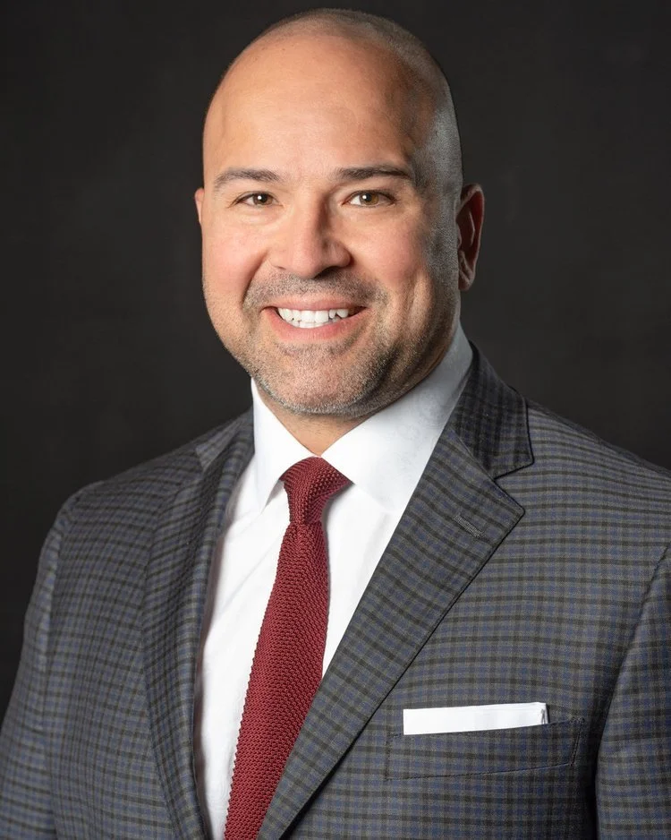 A headshot of a smiling man wearing a checkered suit, white shirt, and red tie, against a dark background.