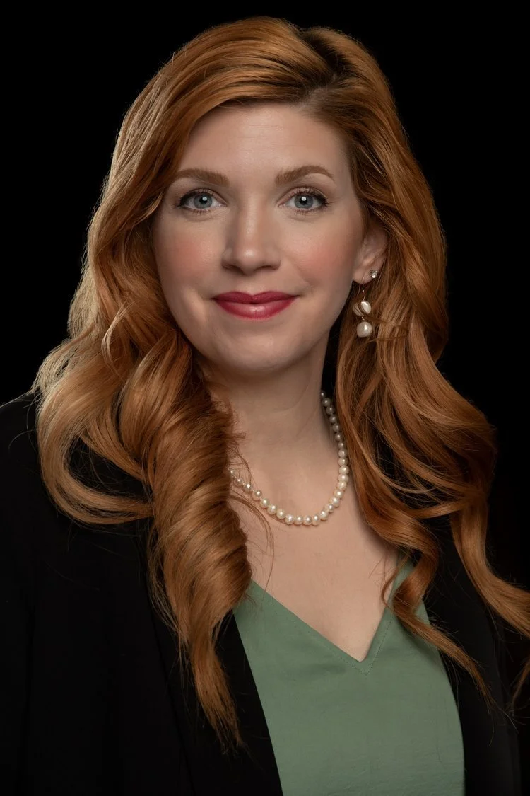 Portrait of a woman with long, wavy red hair, blue eyes, wearing pearl earrings and necklace, a green top, and a black blazer against a black background.