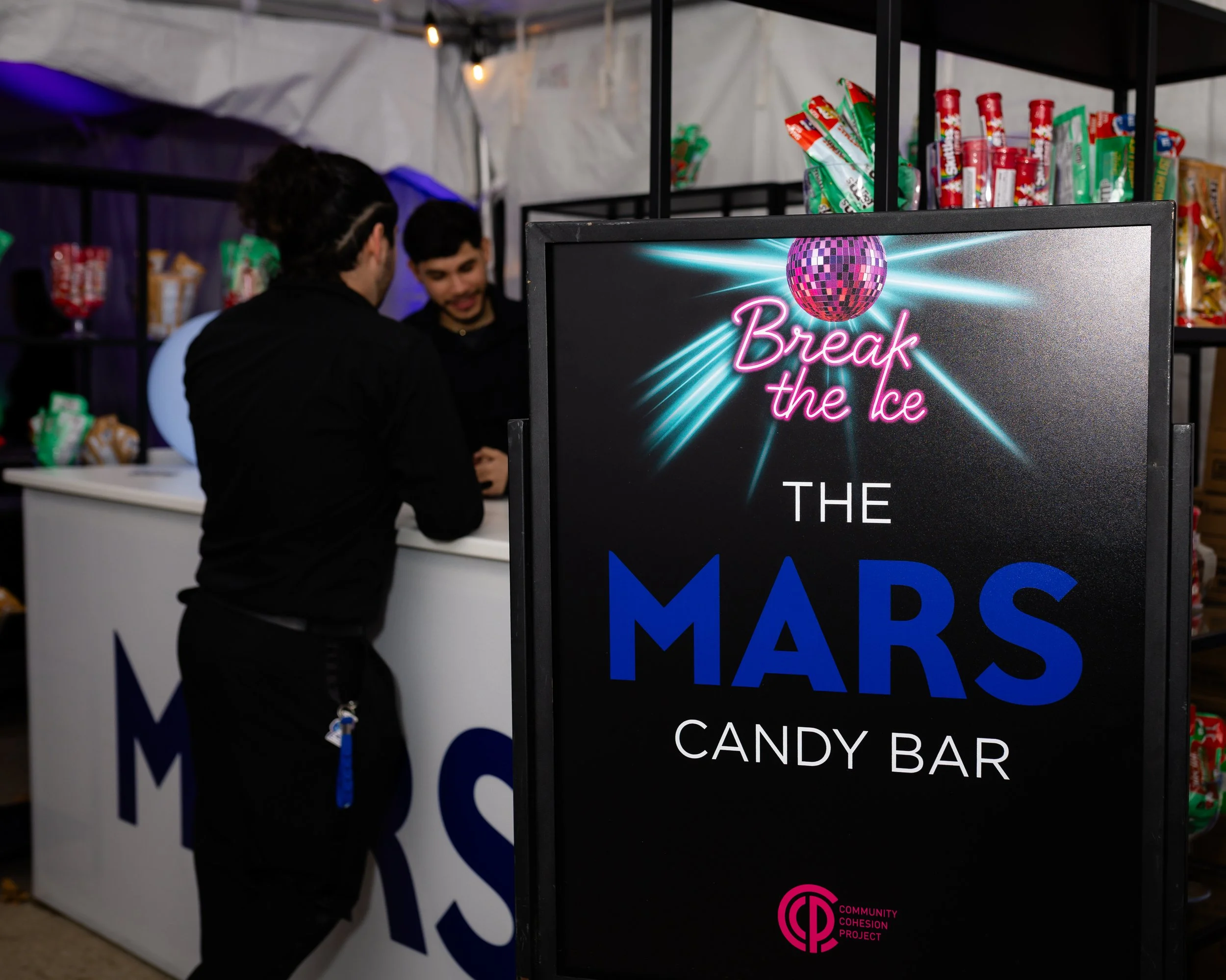 A sign for the Mars Candy Bar at an event, with a neon disco ball and the phrase 'Break the Ice' in bright pink. Two people are talking at a booth in the background, with shelves of candy behind them.