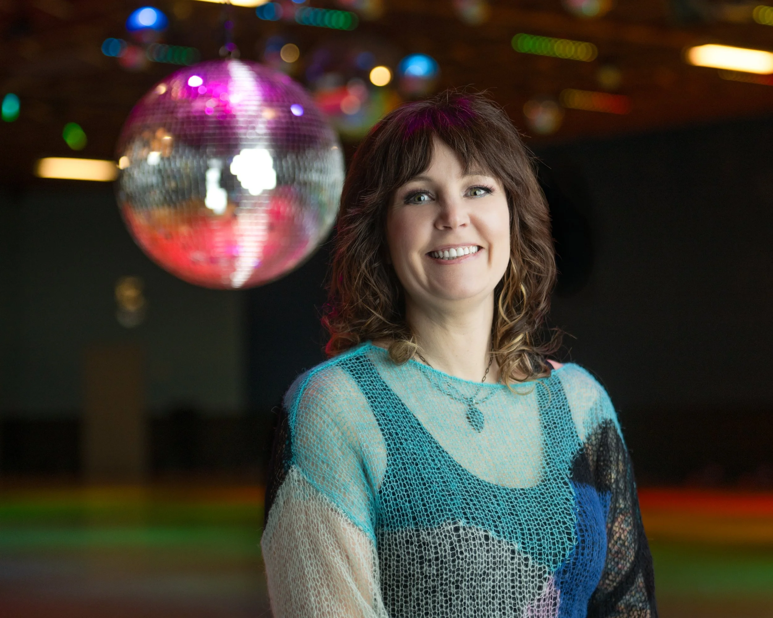 A woman with curly hair smiling in front of a disco ball, in a room with colorful lights.