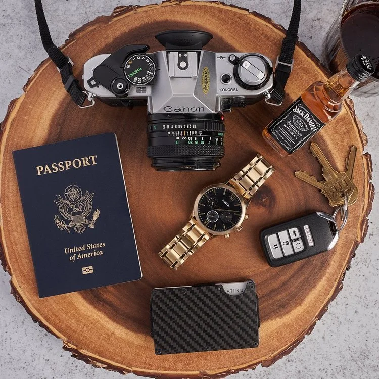 A collection of personal items on a wooden surface, including a Canon camera, a US passport, a gold wristwatch, a set of car keys, a small bottle of Jack Daniel's whiskey, a black wallet, and a gold pen, arranged neatly in a circular pattern.