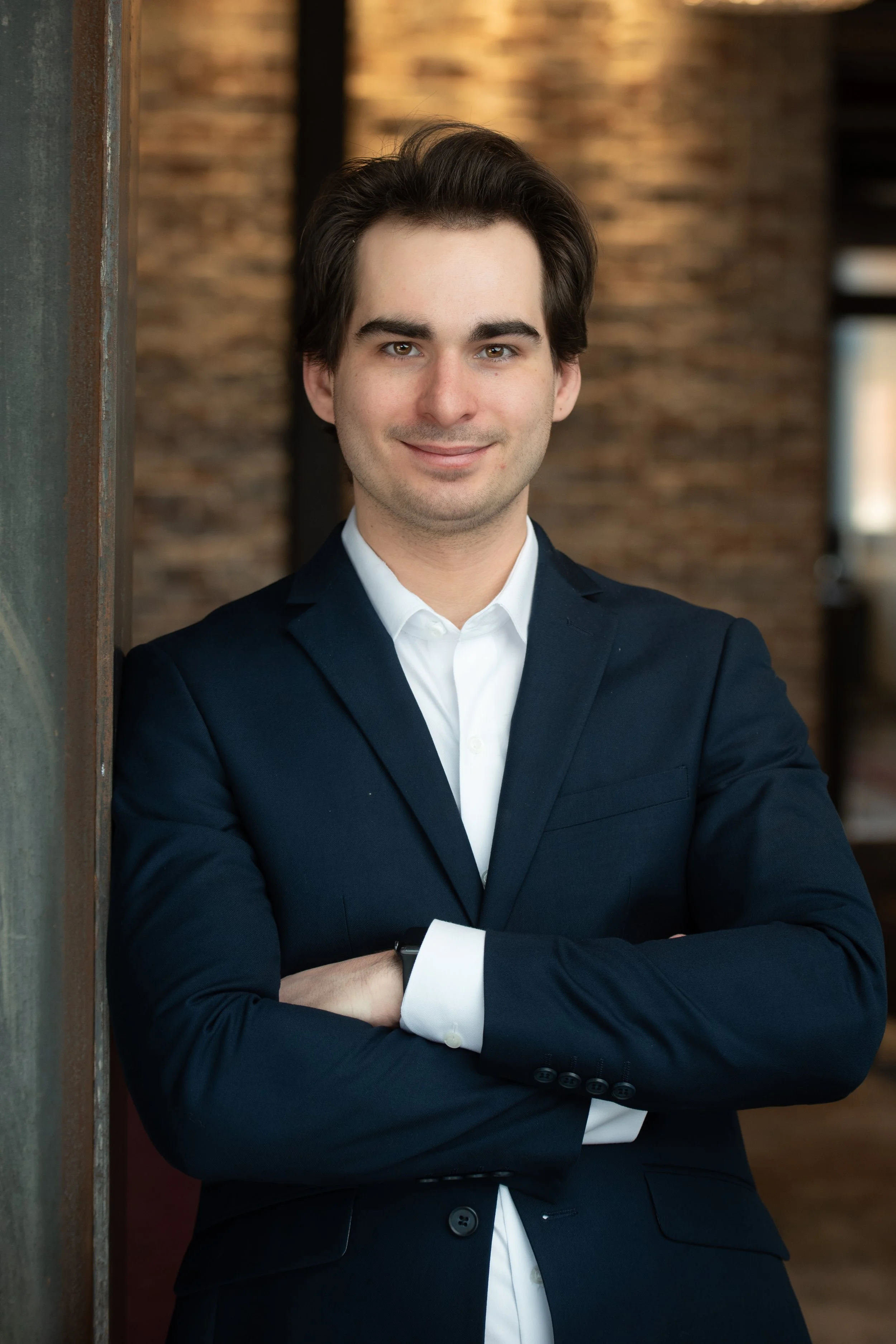 Portrait of a young man in a navy suit with arms crossed, standing indoors against a brick wall. Team headshots of First National Bank of Northwest Arkansas, Fayetteville location.