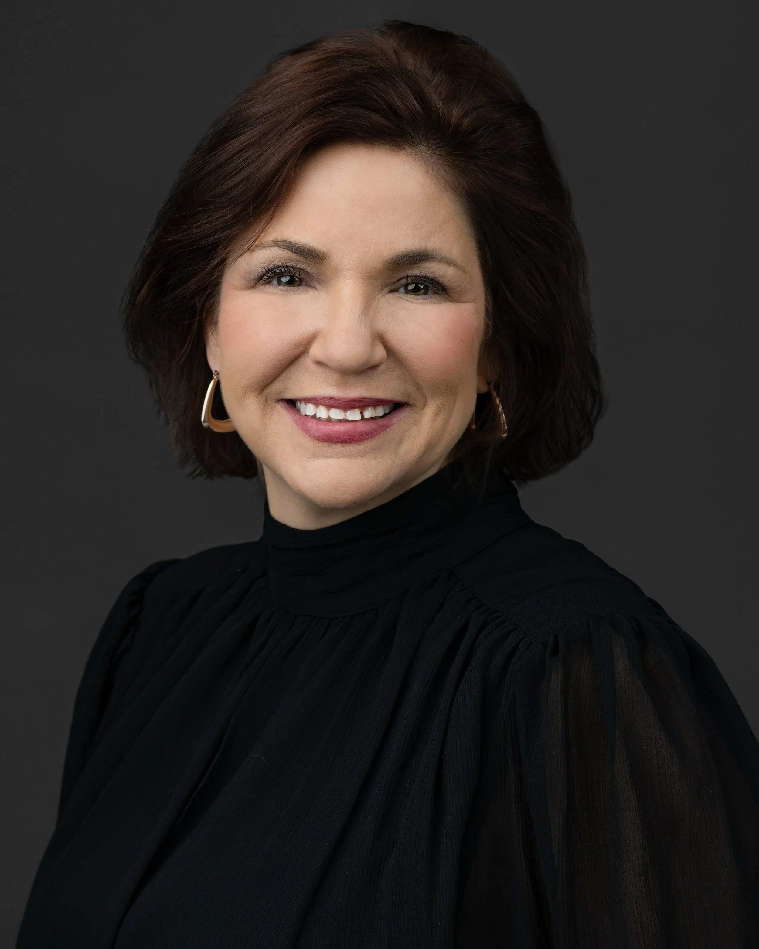 Professional portrait of a smiling woman with dark brown hair wearing a black high-neck blouse and gold hoop earrings against a dark background.