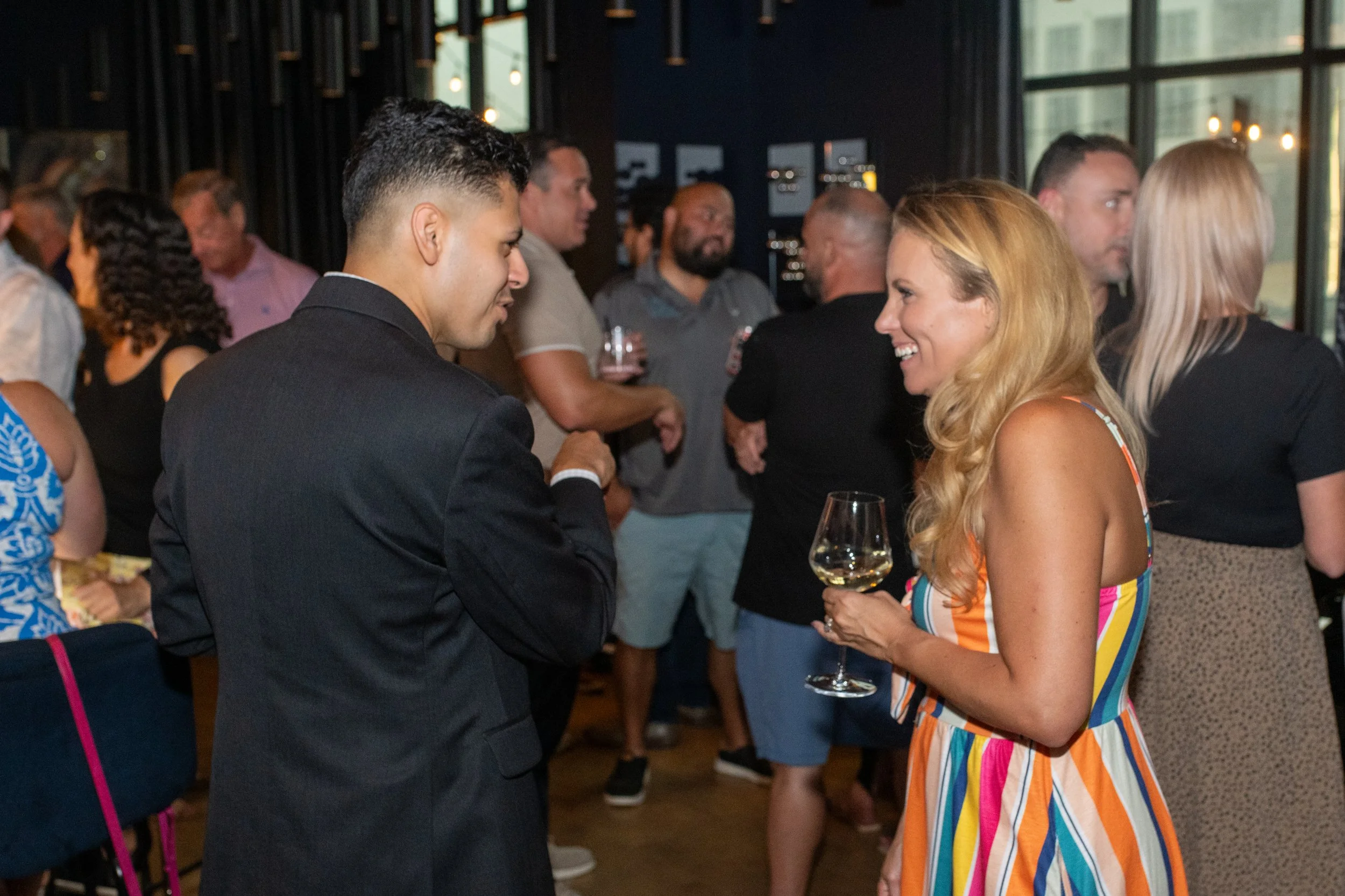 A man in a black suit and a woman in a colorful striped dress are talking and smiling at each other at a social gathering. The woman holds a wine glass, and there are several people in the background mingling and holding drinks in a modern indoor ven