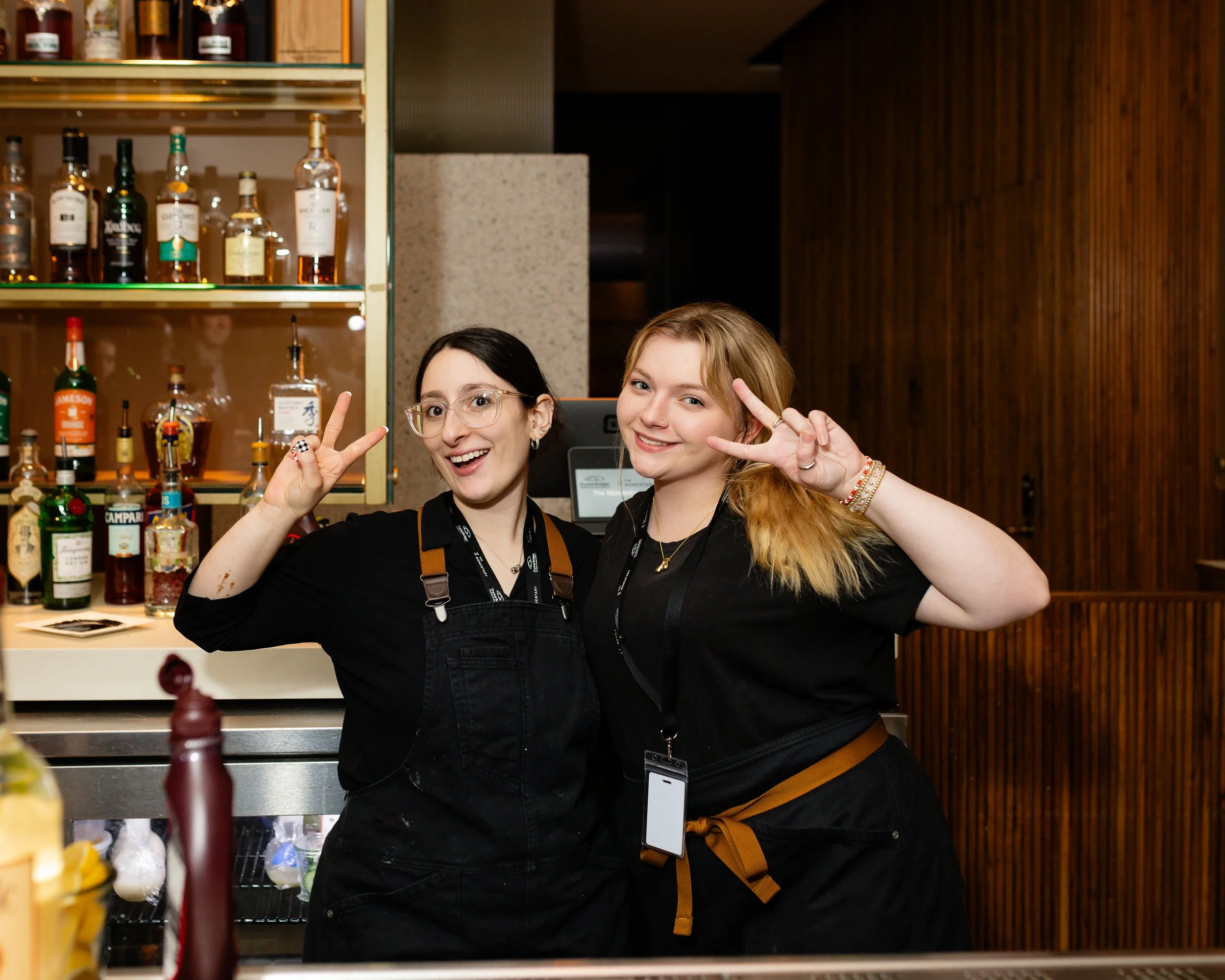 Two smiling women standing behind a bar, making peace signs, with bottles of alcohol on shelves behind them.