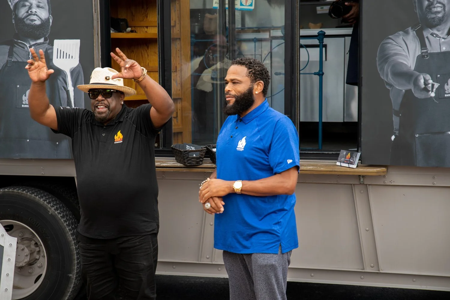 Cedric the Entertainer and Anthony Anderson showcase their AC Barbeque brand at a Walmart Shareholders event in Springdale, Arkansas, standing before a BBQ food truck with cooking posters in the background.