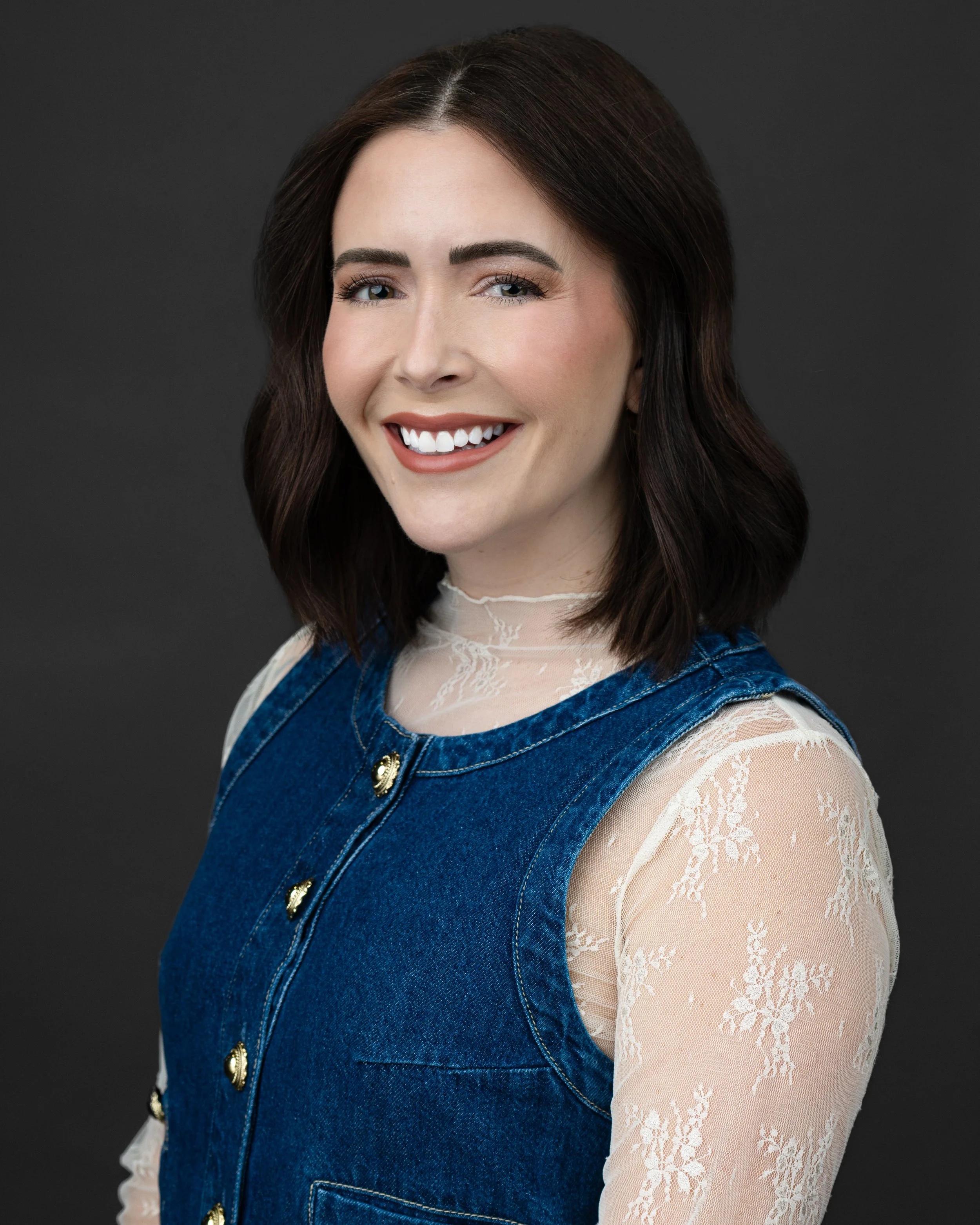 A woman with shoulder-length dark brown hair smiling, wearing a blue denim sleeveless dress with decorative gold buttons and a sheer lace top underneath, against a dark background.