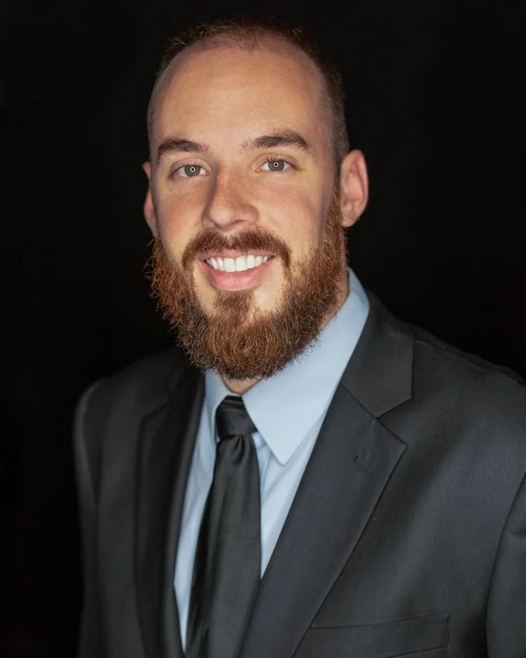 Portrait of a man with a beard in a black suit, light blue shirt, and black tie, smiling against a black background.