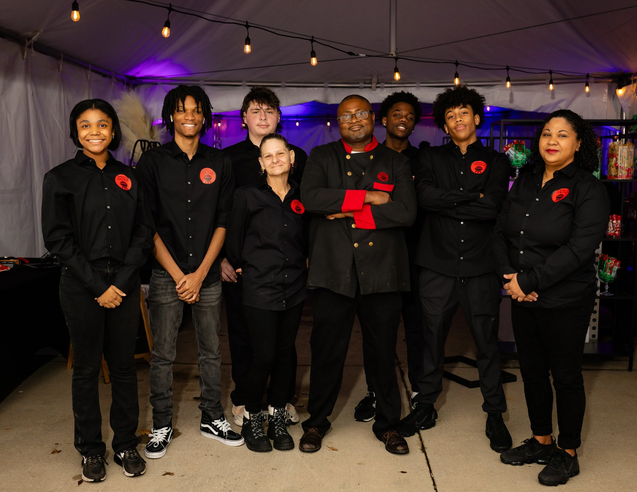 Group of seven people standing together inside a decorated event tent, some with arms crossed, wearing black shirts with red logos, and the man in the center in a black chef's coat with red accents. CEO and Founder Team Nate Walls, Second Hand Smoke.