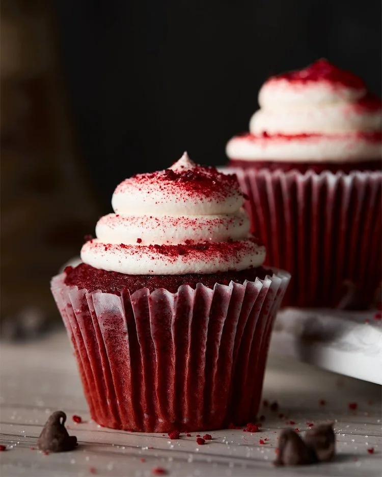 Red velvet cupcake with white frosting and red sprinkles, on a white surface with scattered red crumbs and chocolate chips.