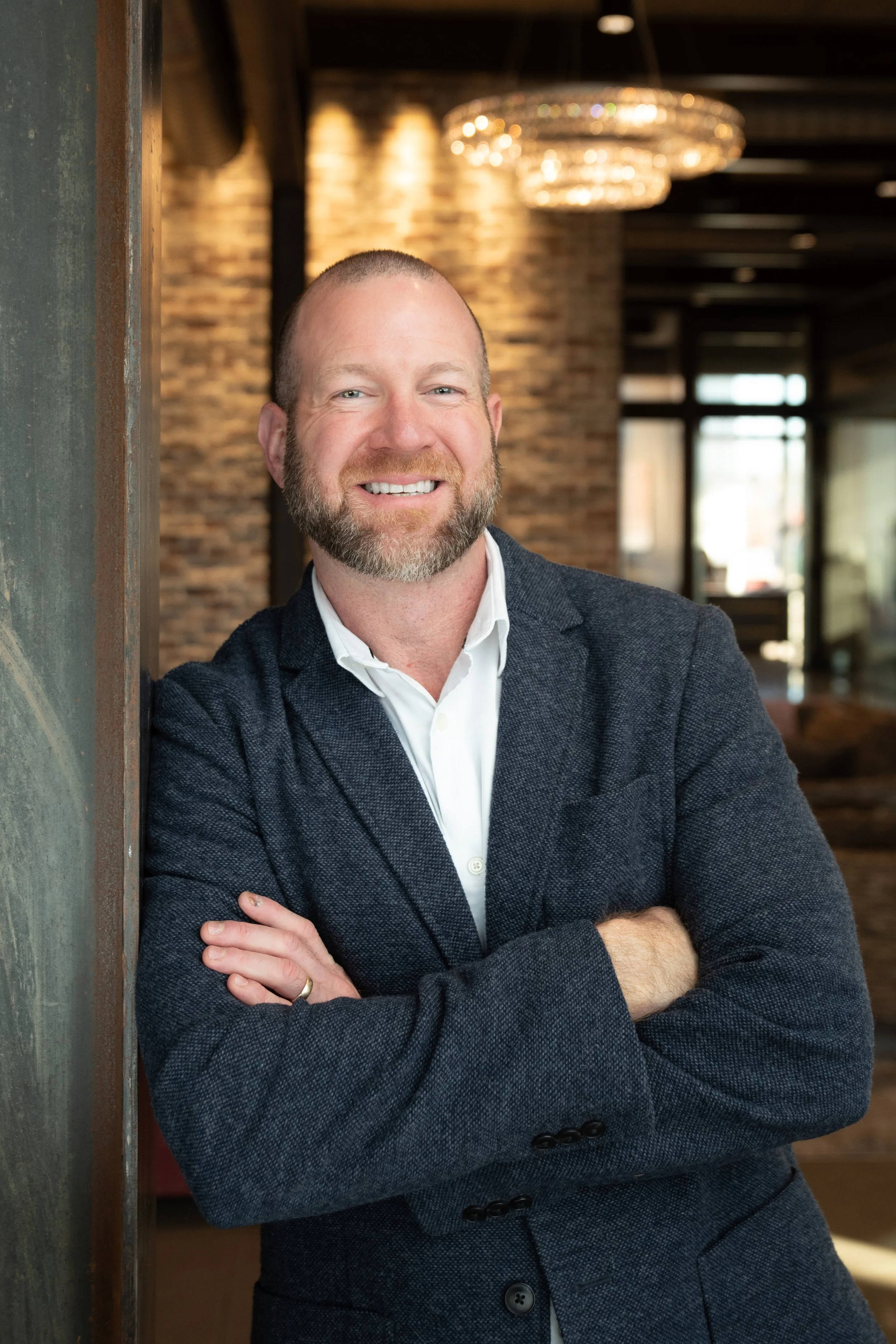 A smiling man with a beard in business attire, crossing his arms, leaning against a wall in a modern, warmly lit interior. Team headshots for First National Bank of Northwest Arkansas, Fayetteville location.