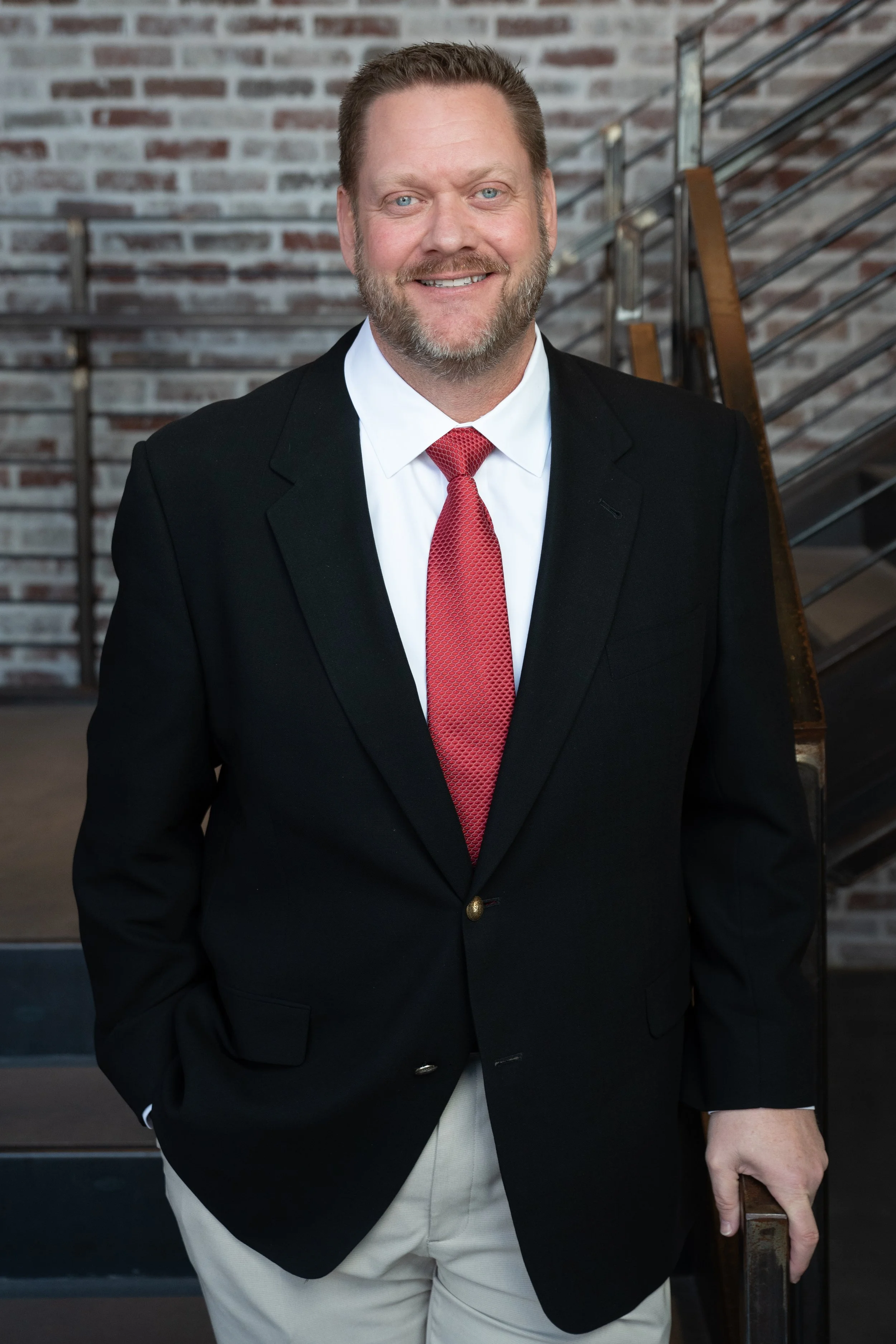 A man wearing a black suit jacket, white shirt, red tie, and light-colored pants, standing indoors near a staircase with a brick wall background. President of First National Bank of Northwest Arkansas, Bentonville location.