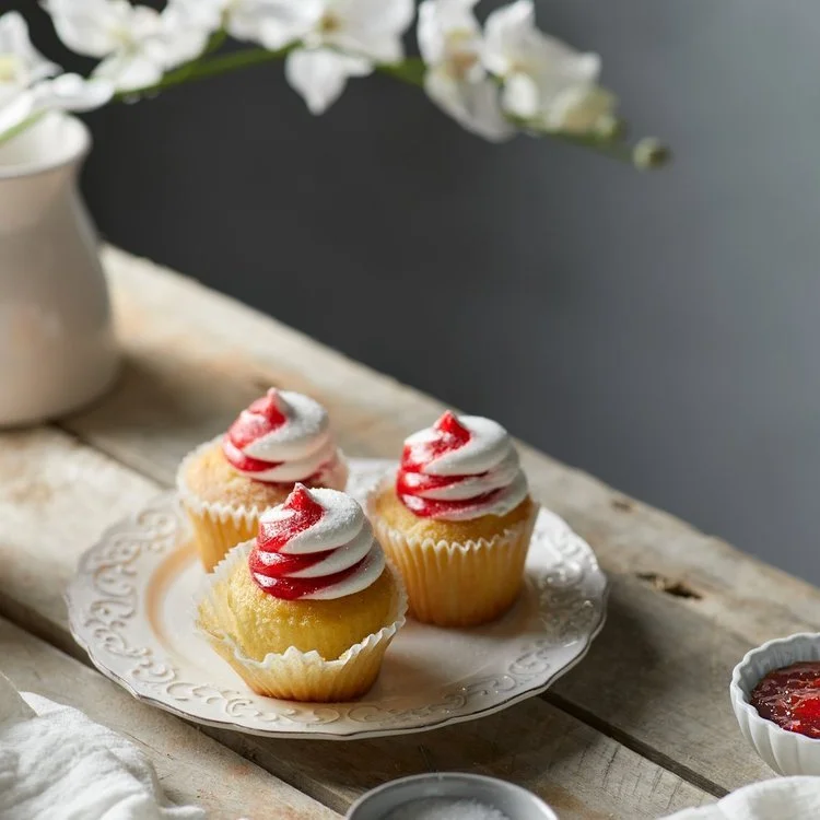 Four cupcakes with white and red swirled frosting on a decorative white plate, placed on a rustic wooden table.