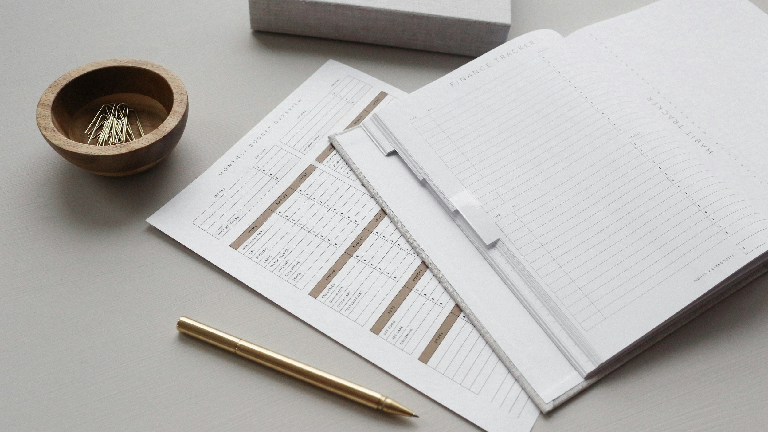 Office desk with financial planning documents, a gold pen, a small wooden bowl with paper clips, and a gray binder.