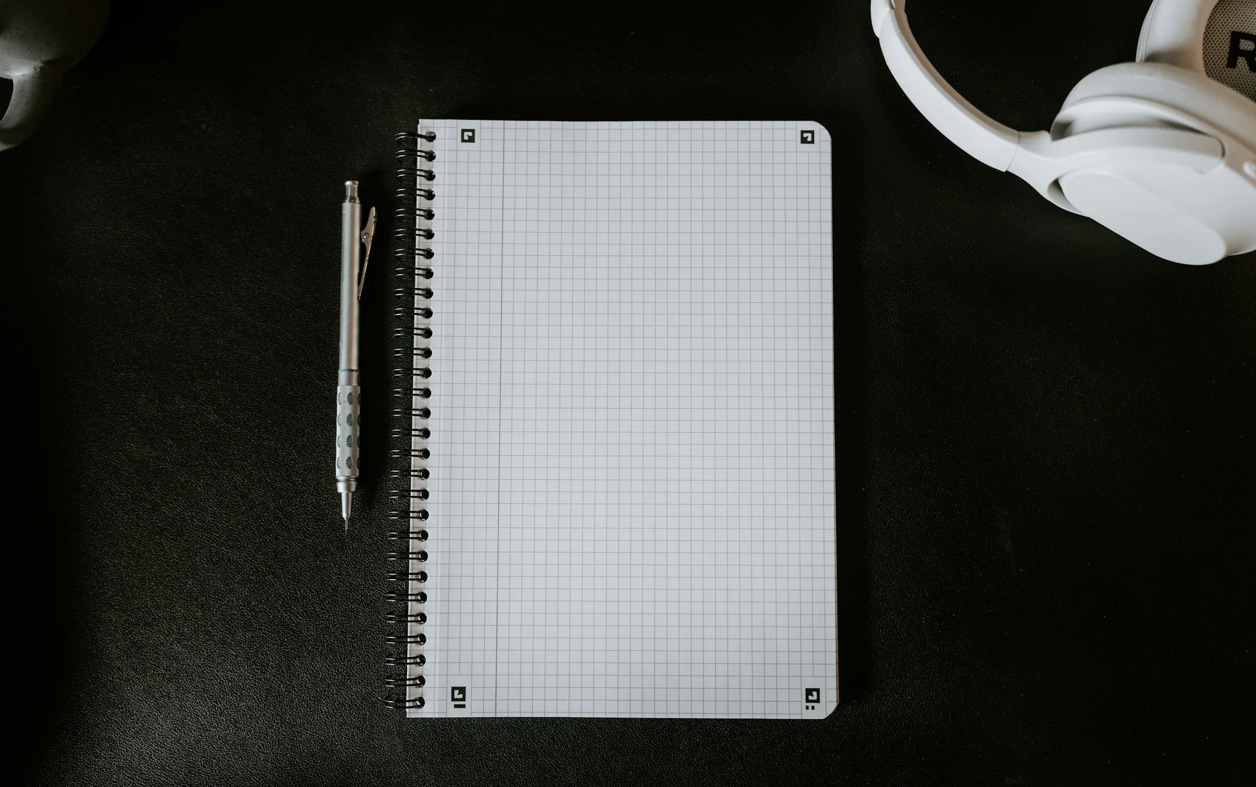 A black desk with a spiral notebook with graph paper, a silver pen, and white over-ear headphones.