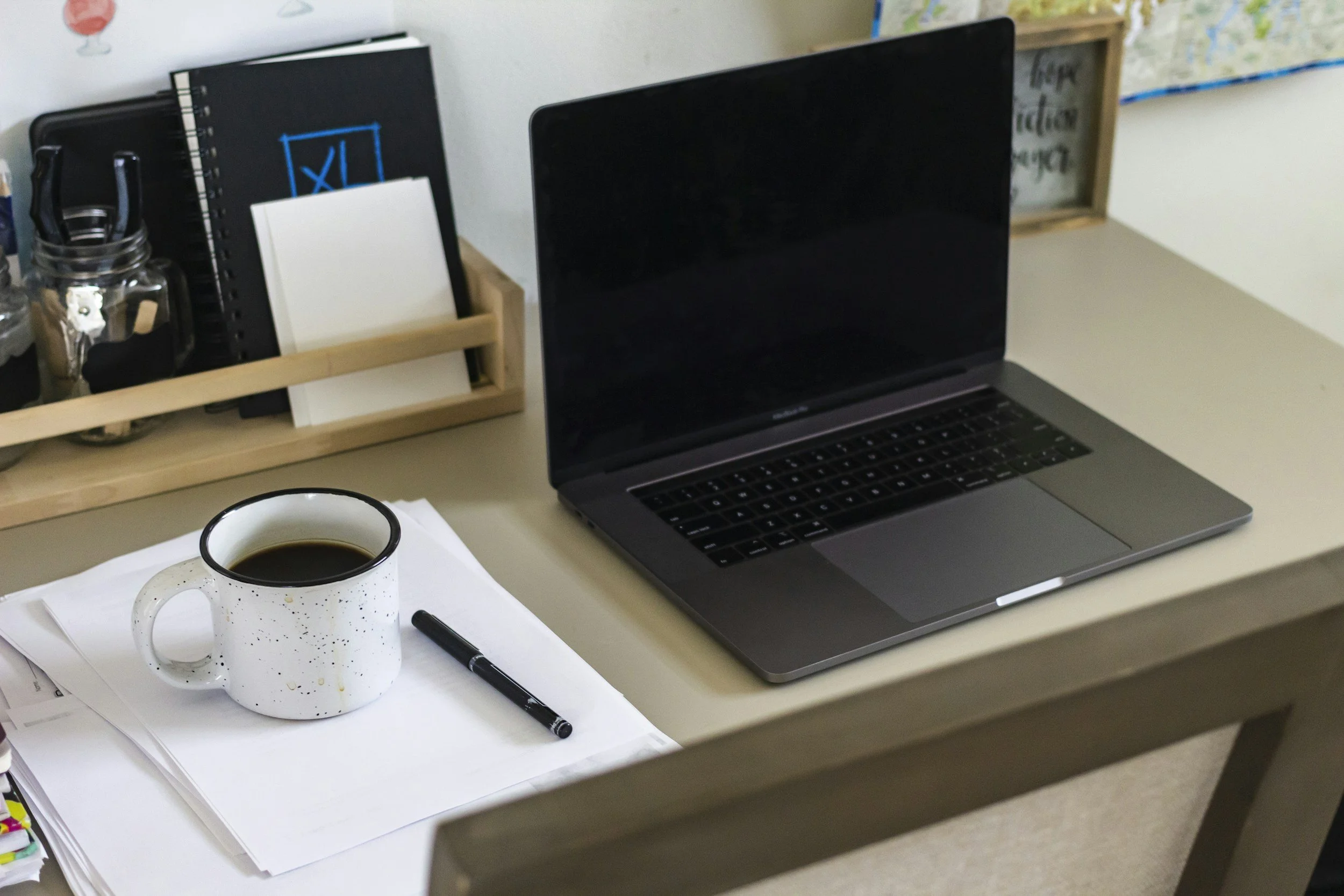 A desk with an open laptop, a white mug filled with coffee, a black pen, papers, and an organizer with notebooks and writing supplies.