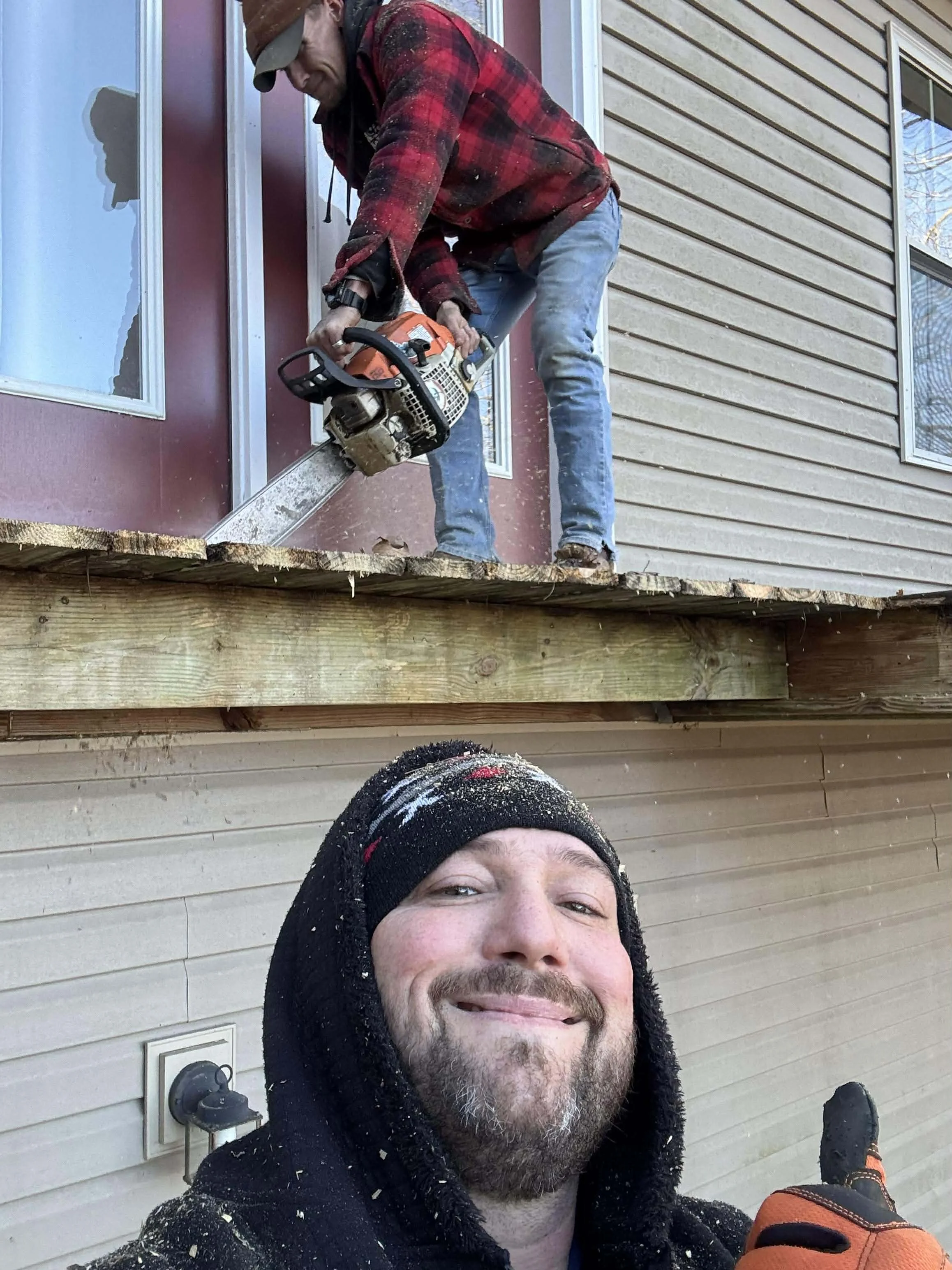 Two men working on house exterior; one using a chainsaw to cut wood on a deck, the other taking a selfie.
