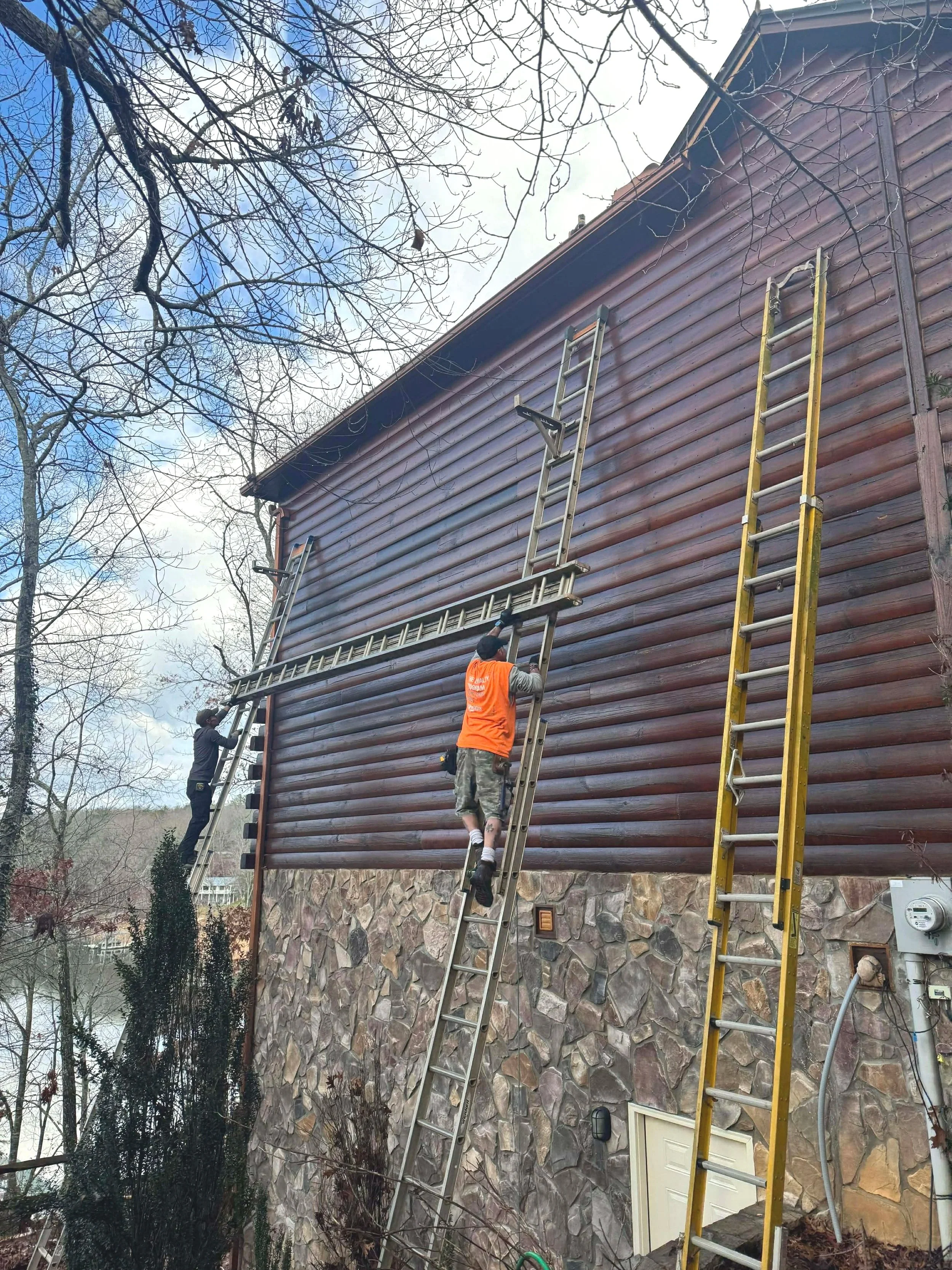 Three workers installing or repairing ladders on a large wooden house, with two ladders leaning against the house and one person working on top of a ladder. The house has a stone foundation and a brown wooden exterior, with bare tree branches and a cloudy sky in the background.
